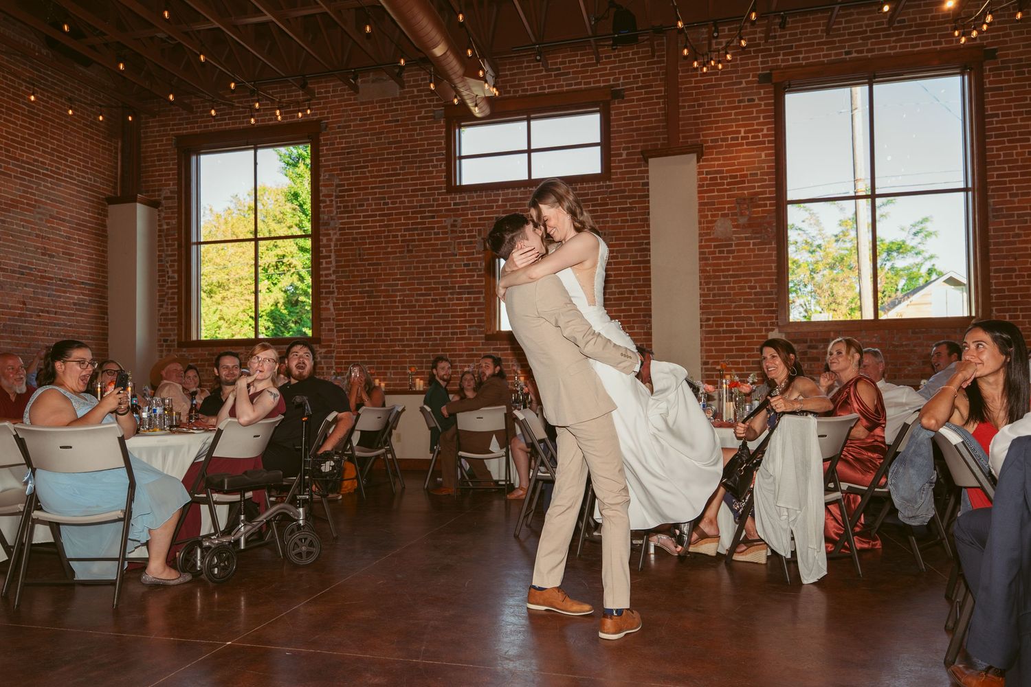 Wedding reception in a rustic brick venue with string lights and large windows.