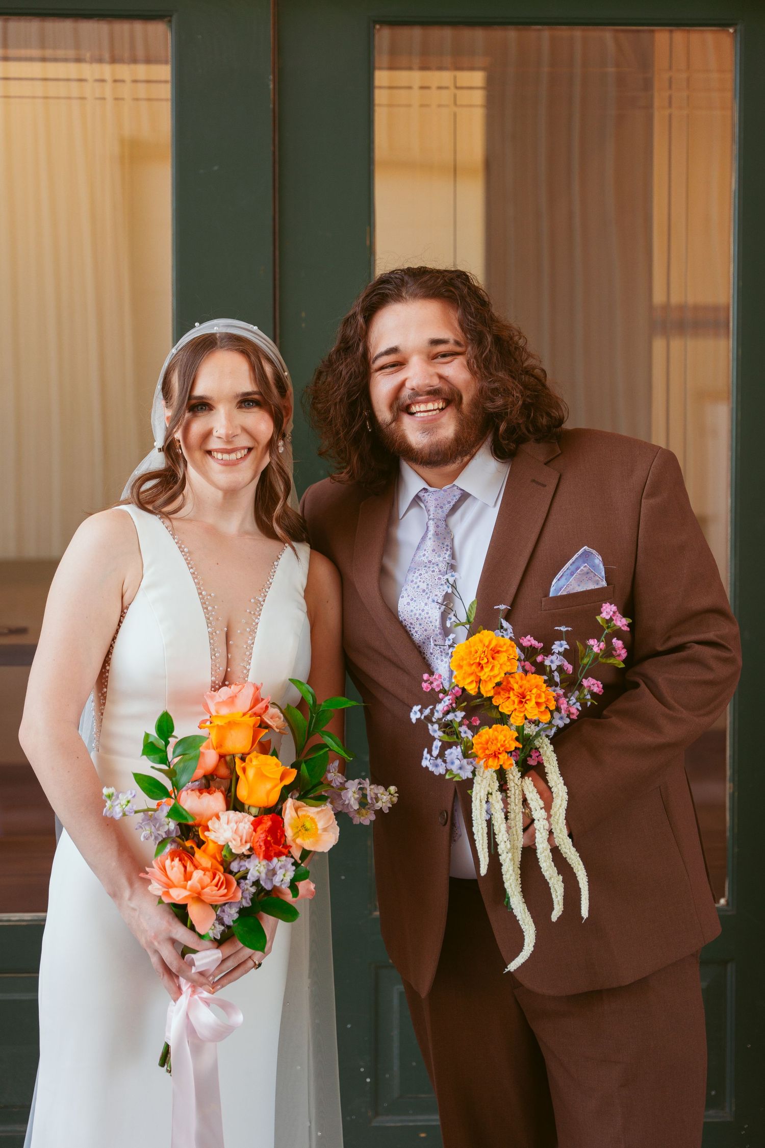 A wedding couple poses together in formal attire with vibrant orange and pink bouquets against a green door.