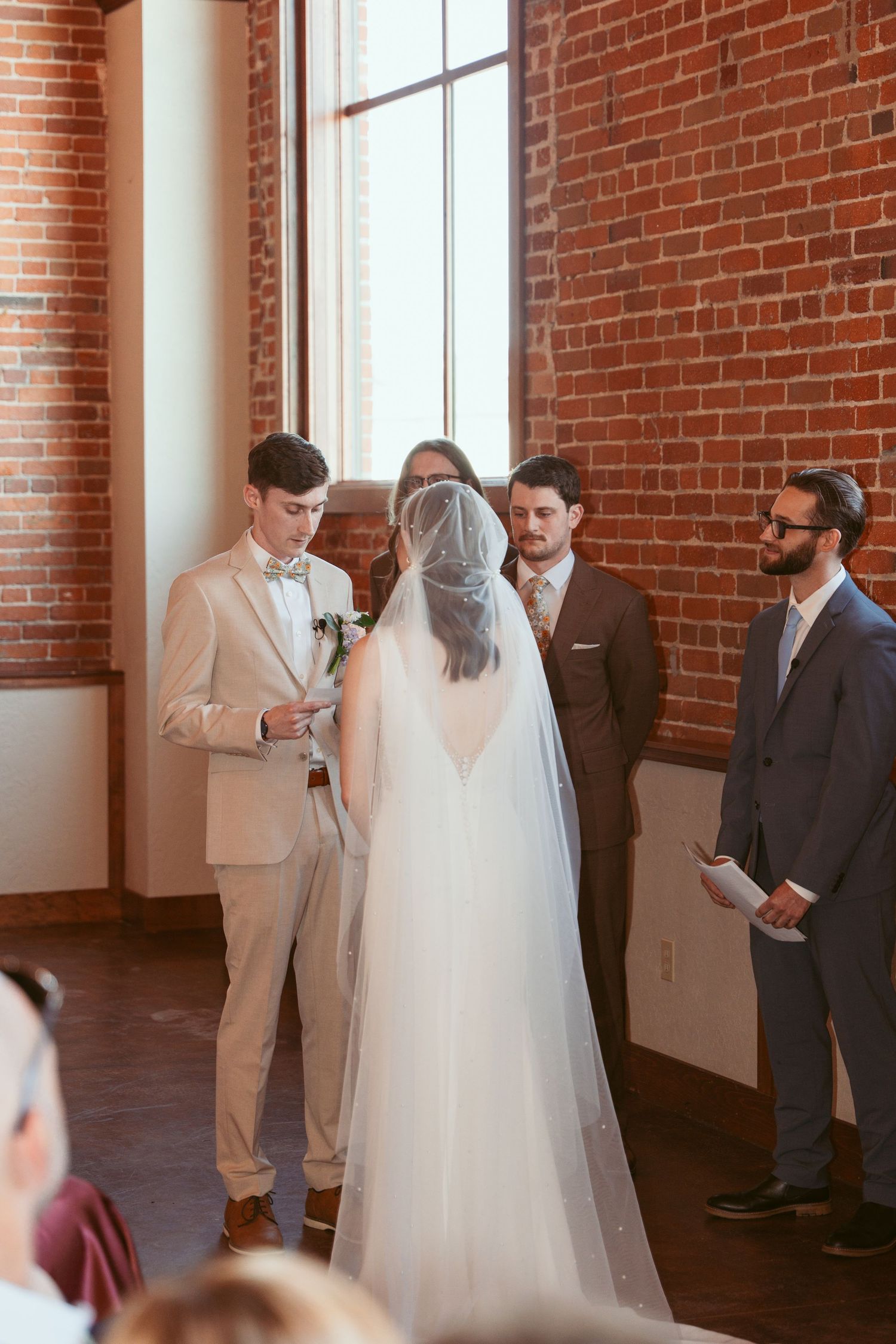 Wedding party gathered for ceremony in rustic brick-walled venue with large windows.
