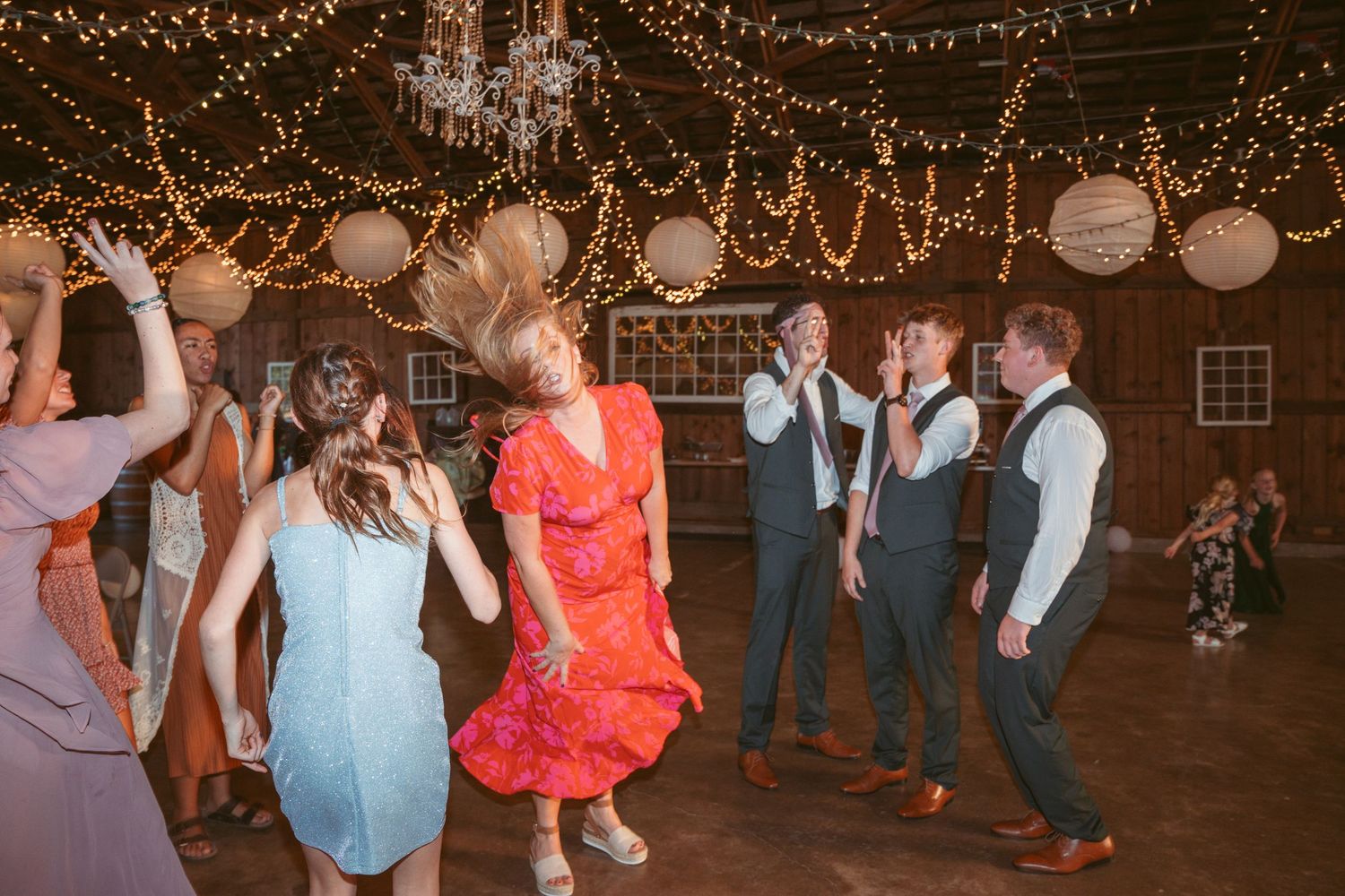 Wedding reception guests dancing under string lights and paper lanterns in a rustic barn setting.