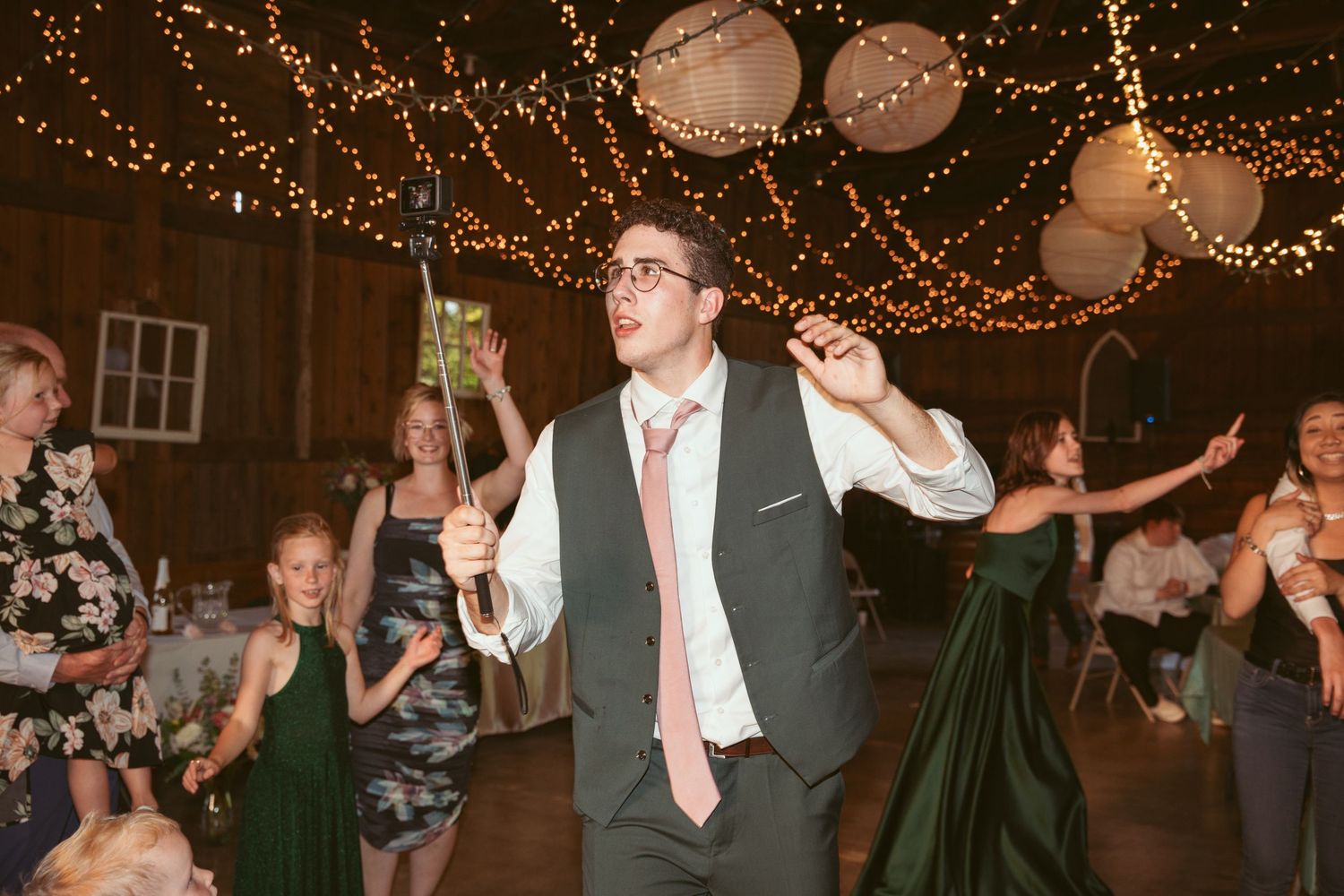 Wedding celebration with guests dancing under twinkle lights in a rustic barn setting.