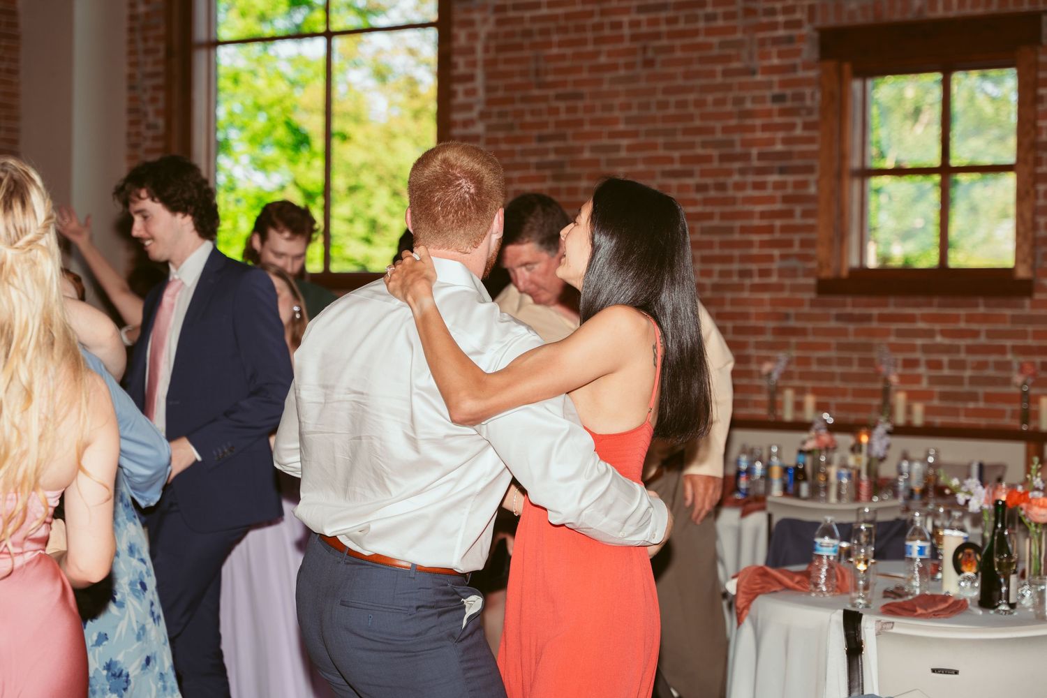 Wedding guests celebrate on the dance floor of a rustic brick venue with large windows.