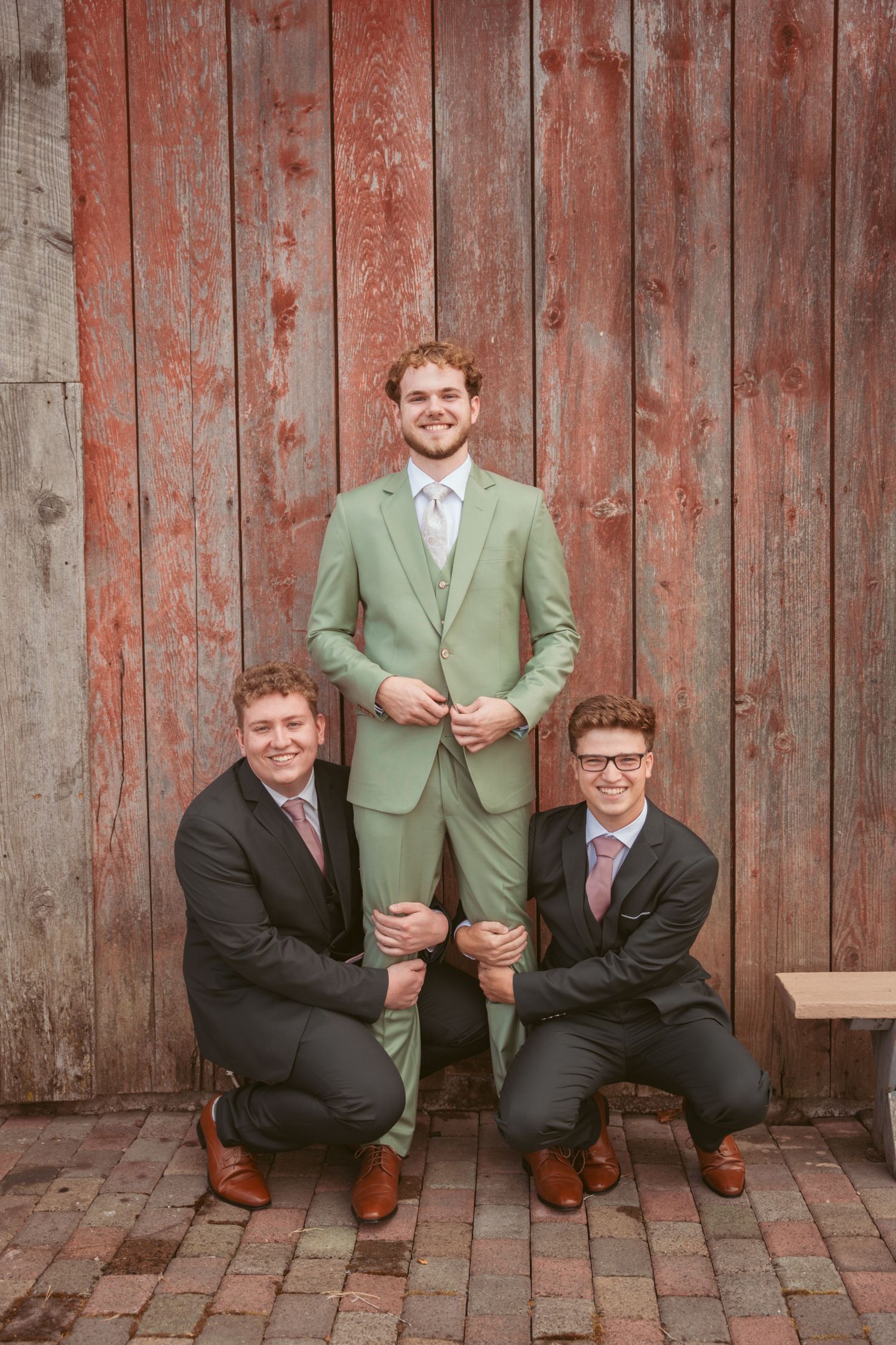 Three groomsmen pose playfully against a rustic red barn wall, with one standing while two kneel beside him.
