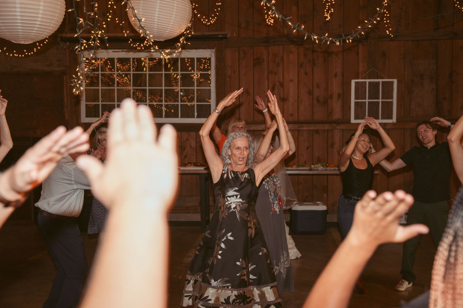 Guests dancing at a barn reception with twinkle lights and lanterns hanging overhead.