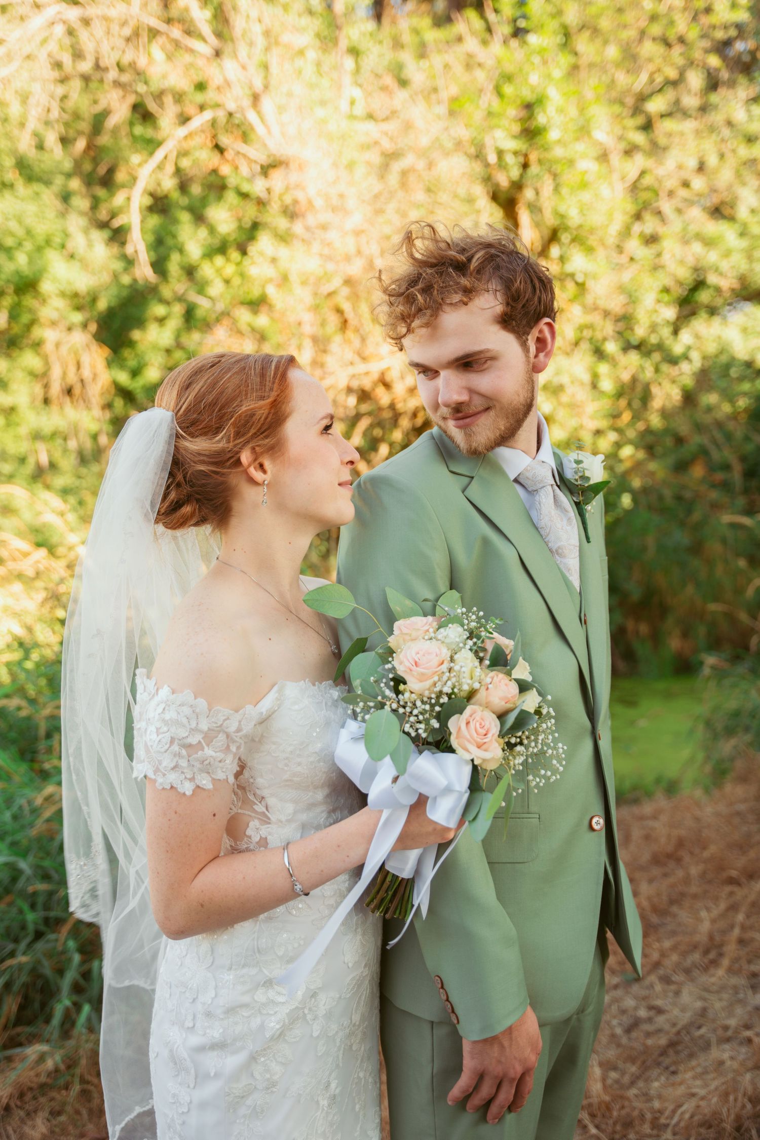 Newlyweds share tender moments in sage green suit and lace wedding dress outdoors.