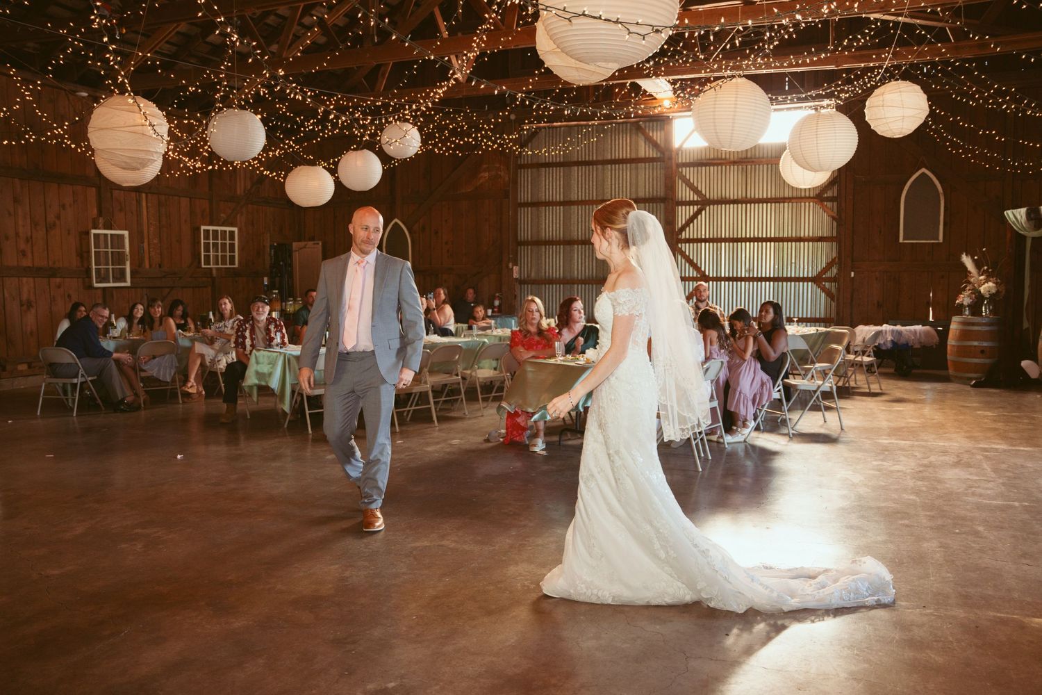 Wedding dance performance in a rustic barn setting with decorative lighting and watching guests.