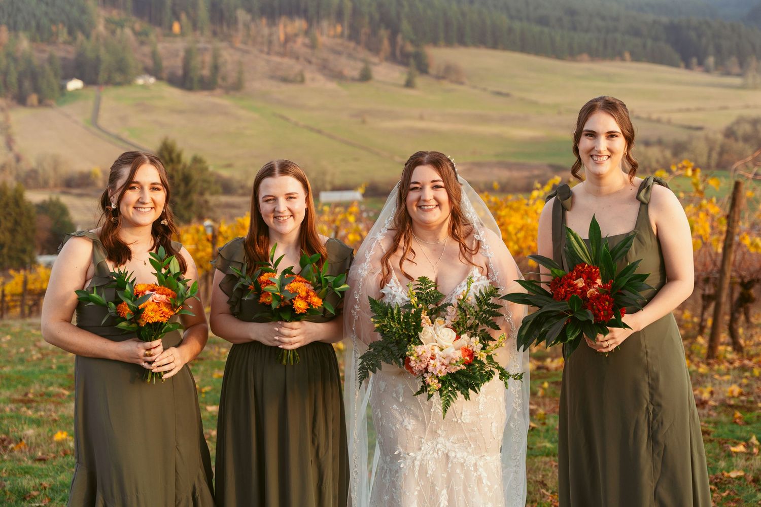 Wedding party wearing olive green dresses poses in front of autumn vineyard landscape at sunset.
