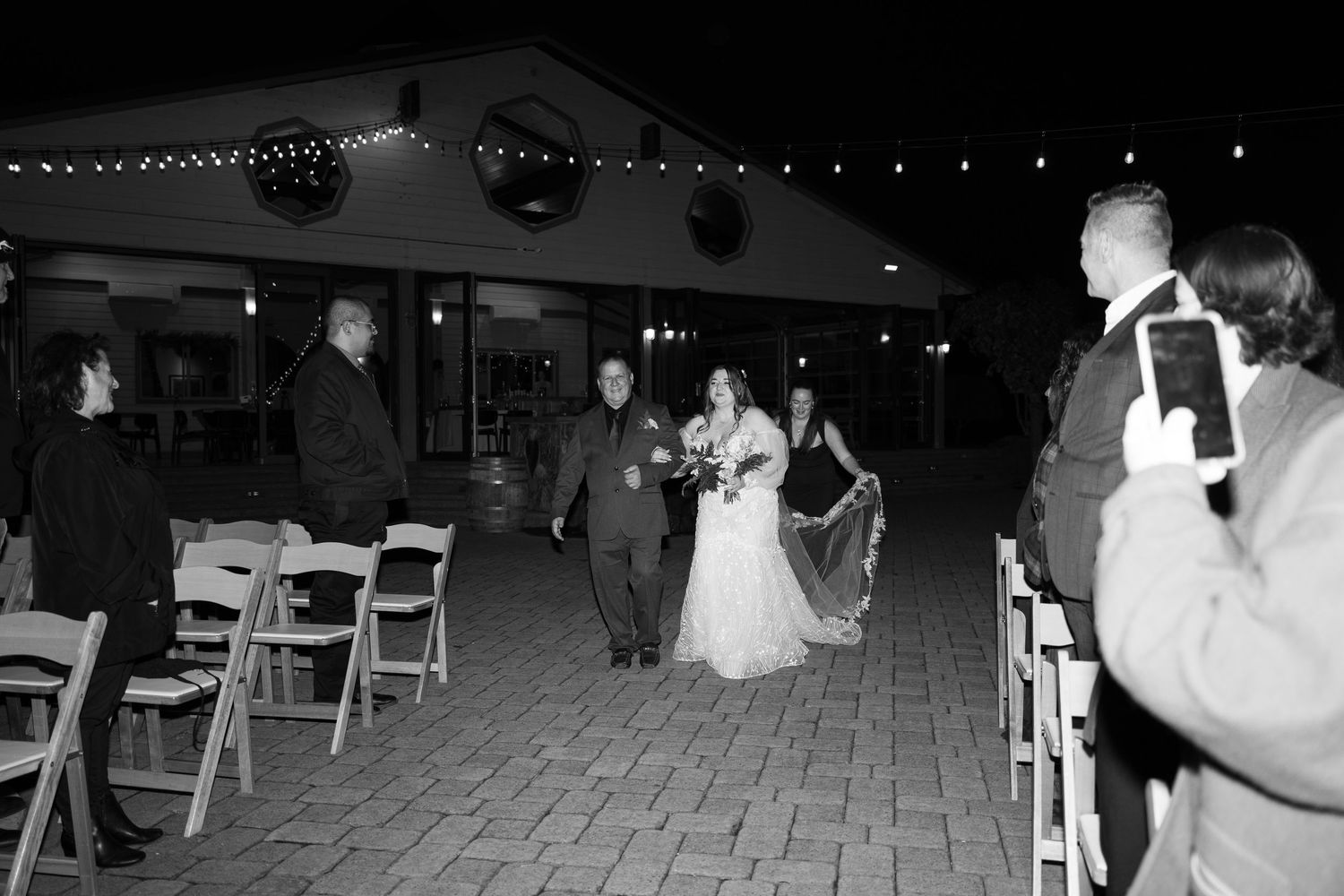 Wedding celebration on a dimly lit stone patio with wooden chairs and string lights overhead.