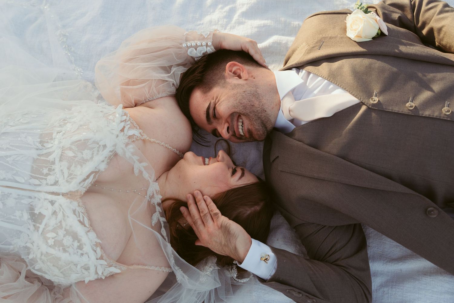 Couple lying together on white blanket in vineyard with mountain landscape behind them.