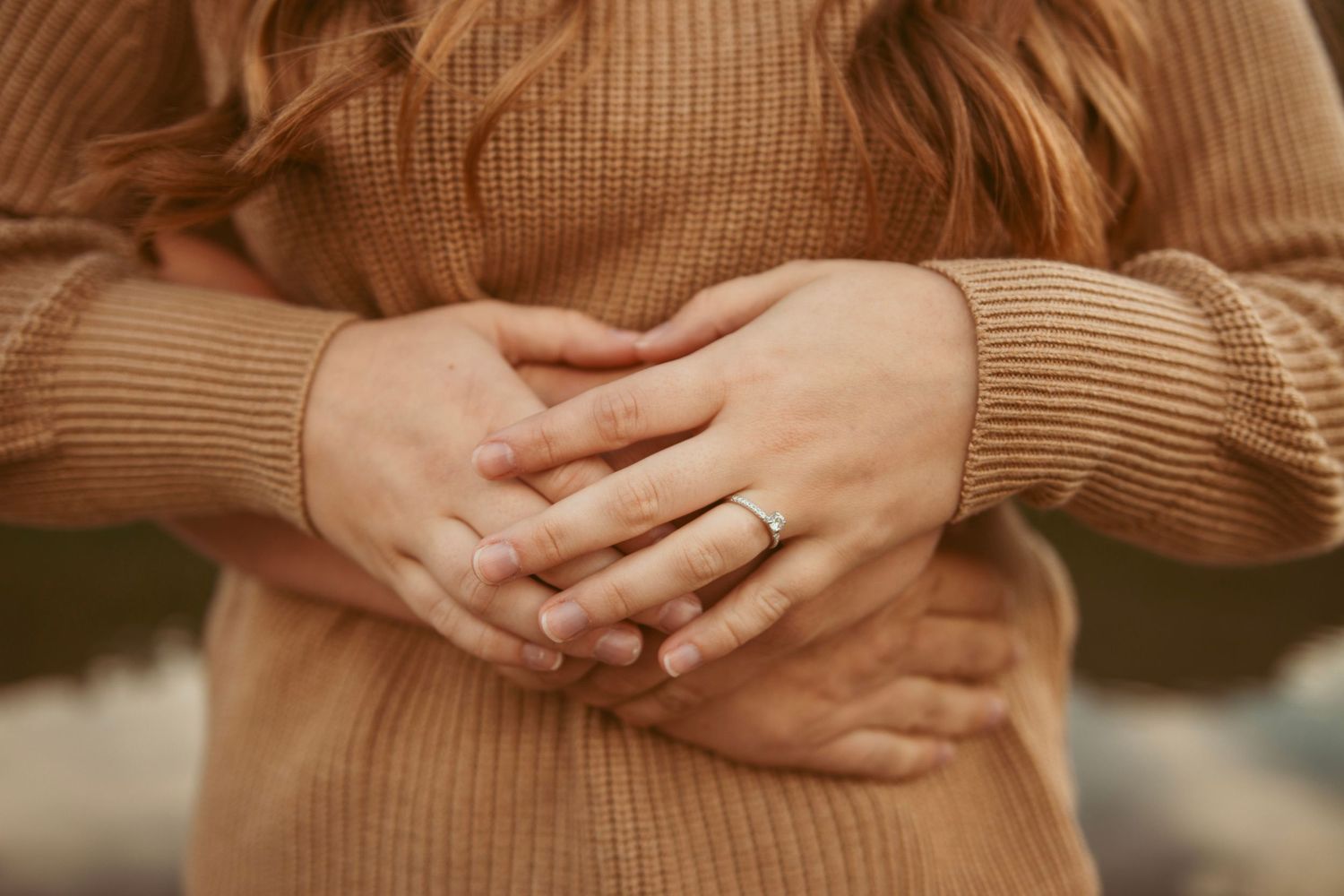 Close-up of hands wearing engagement ring against tan knit sweater.