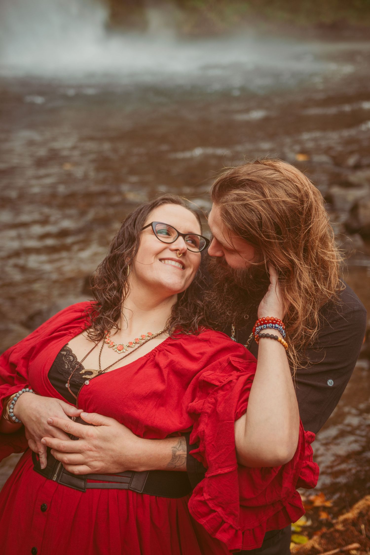 A romantic couple embraces near a waterfall, with one wearing a flowing red dress and the other in dark clothing.