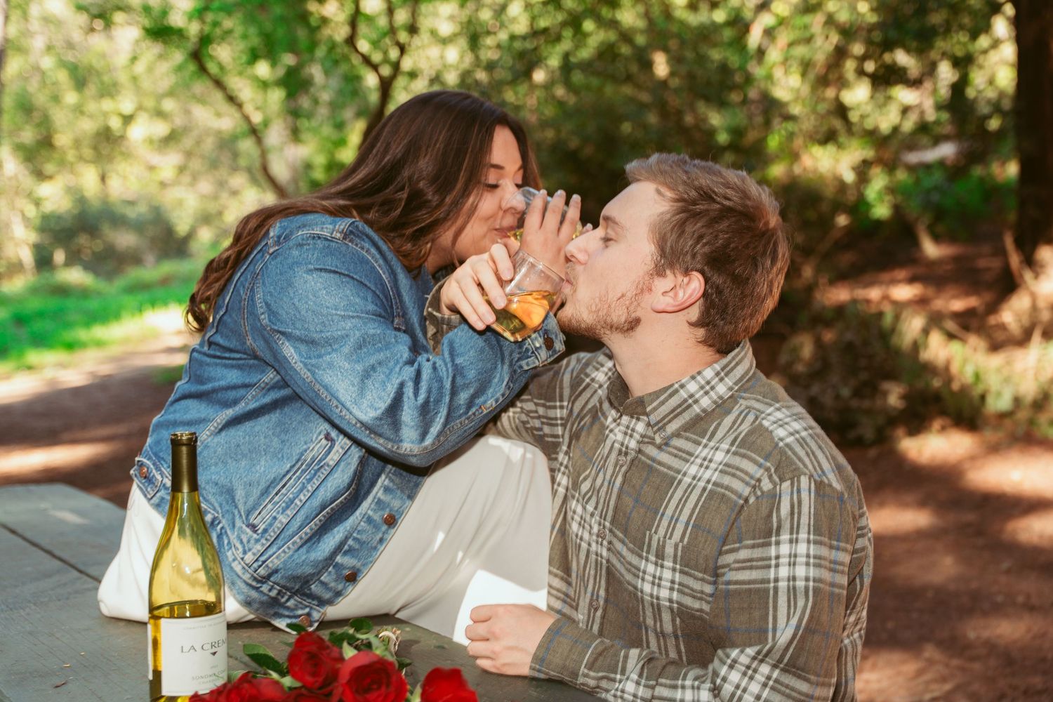A couple shares a romantic moment on a forest bench surrounded by tall trees.