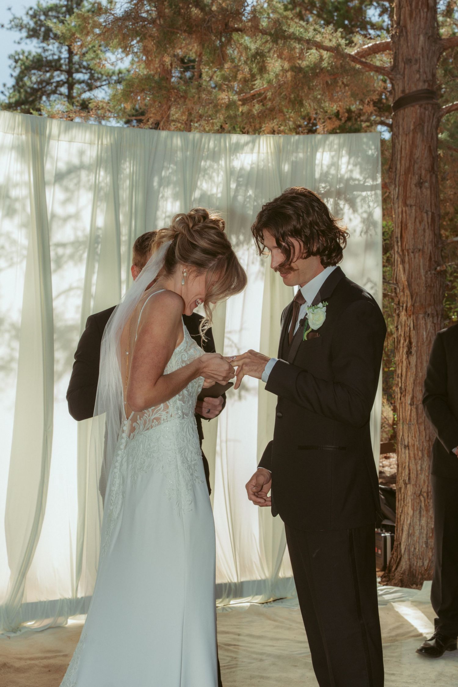 Couple exchanging rings during forest wedding ceremony with white curtain backdrop.