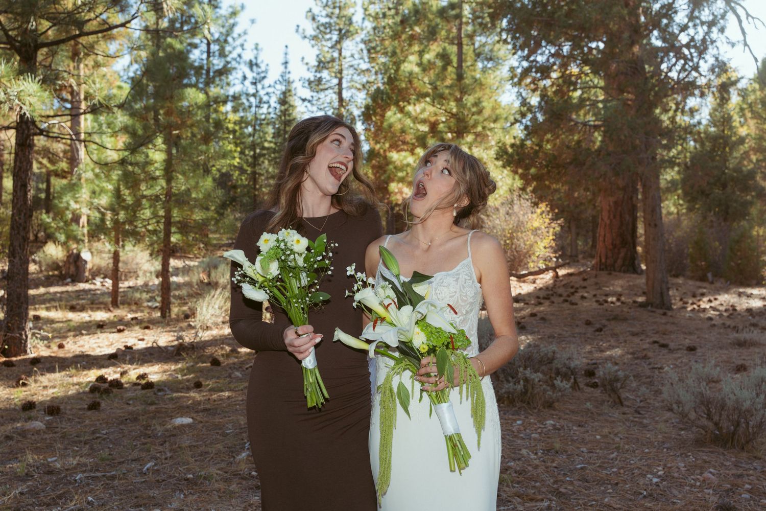 Two people laugh together while holding bouquets in a pine forest setting.
