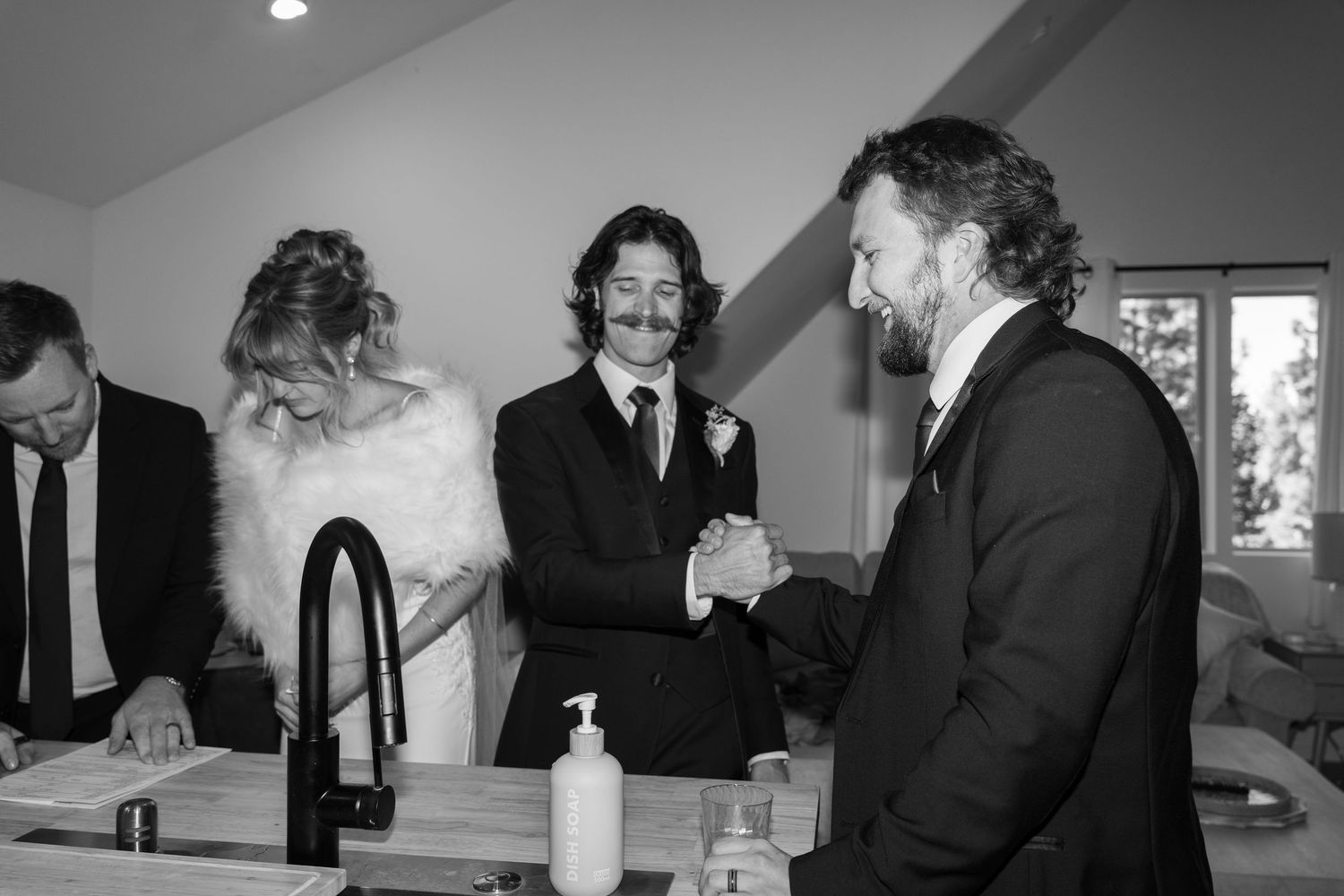 Wedding party members gathered around a kitchen counter in formal attire.