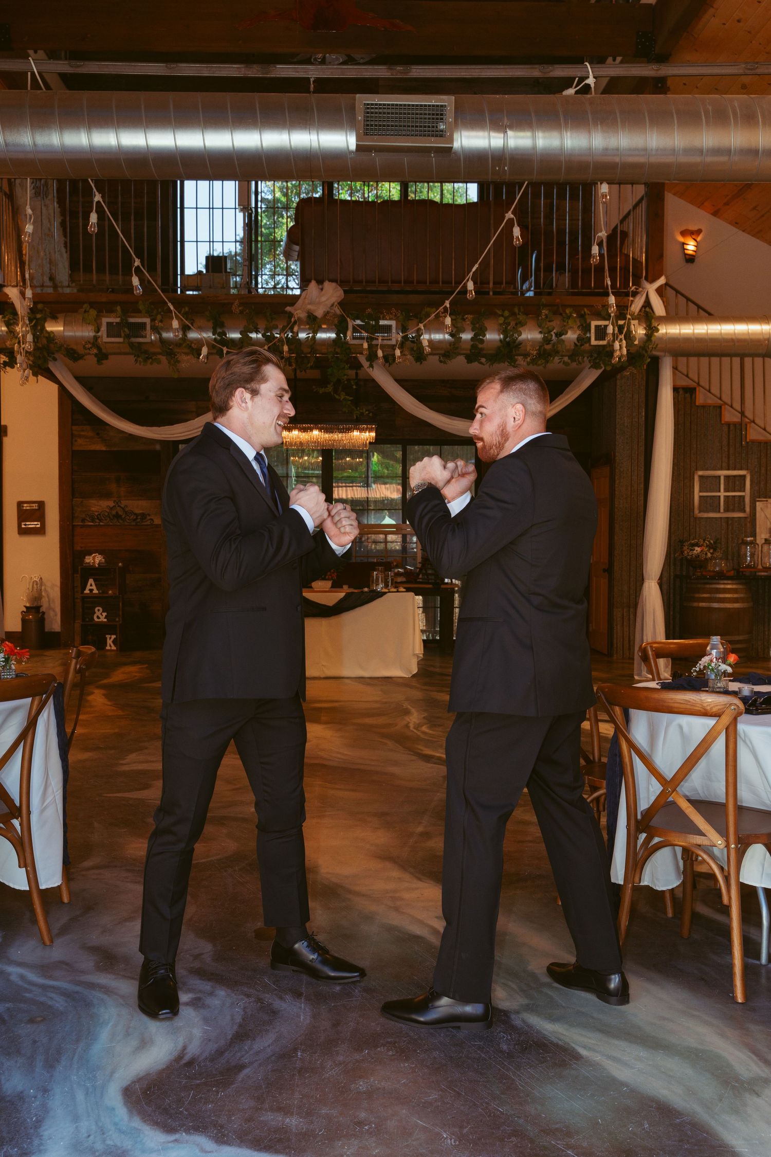 Two men in black suits playfully square off in a rustic indoor venue with exposed ductwork and string lights.