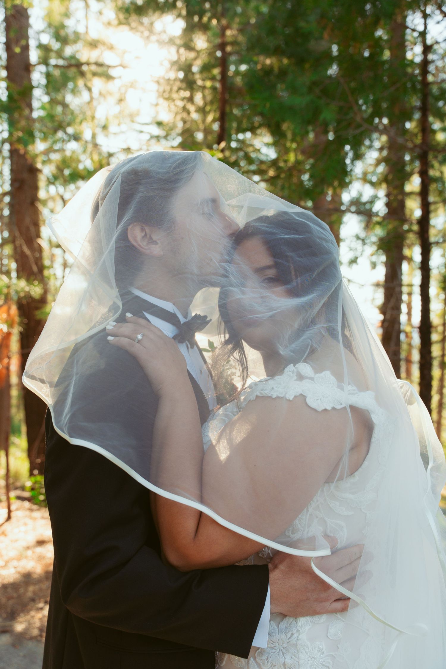 A romantic kiss under a flowing veil in dappled forest sunlight.
