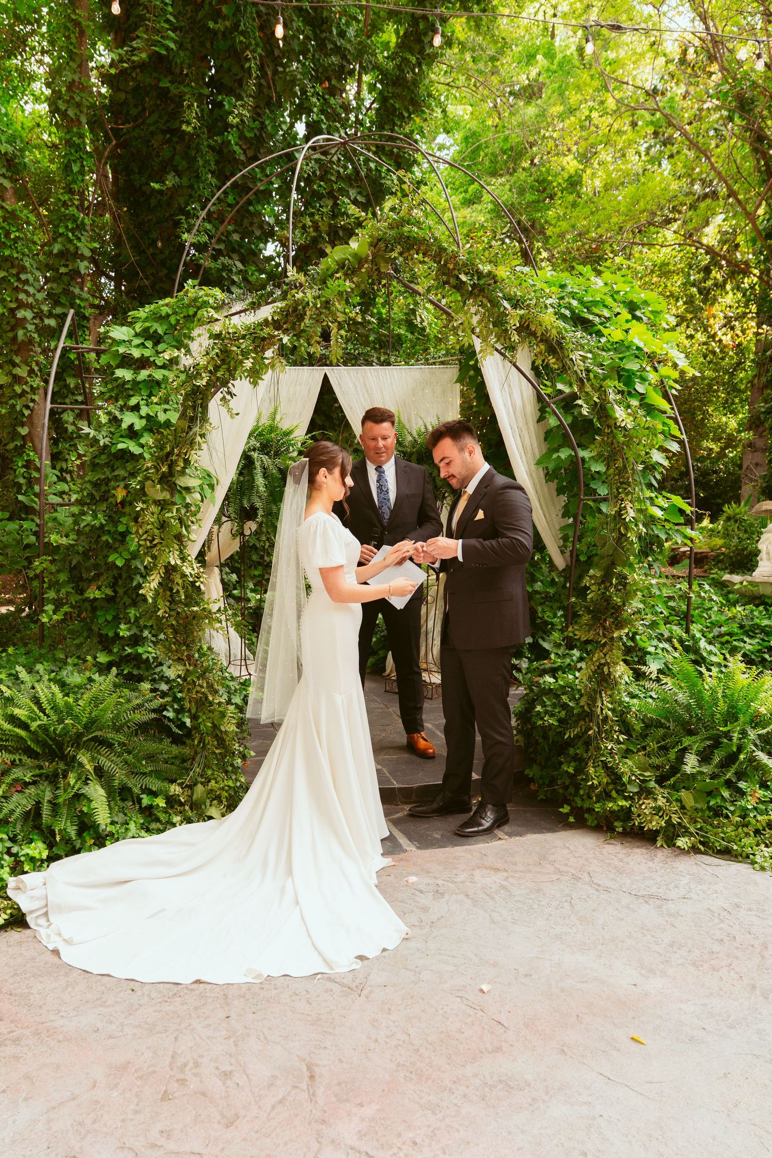 Bride and groom share their first kiss as newlyweds under a garden archway with lush foliage.