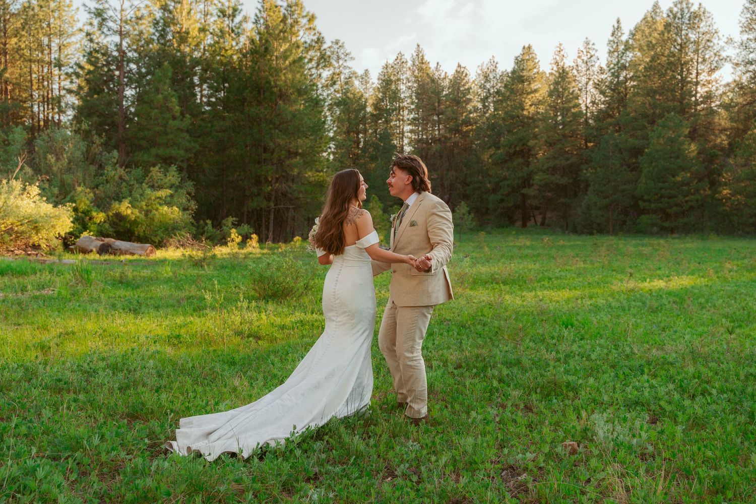 Wedding couple walking hand in hand through a green meadow with pine forest backdrop.