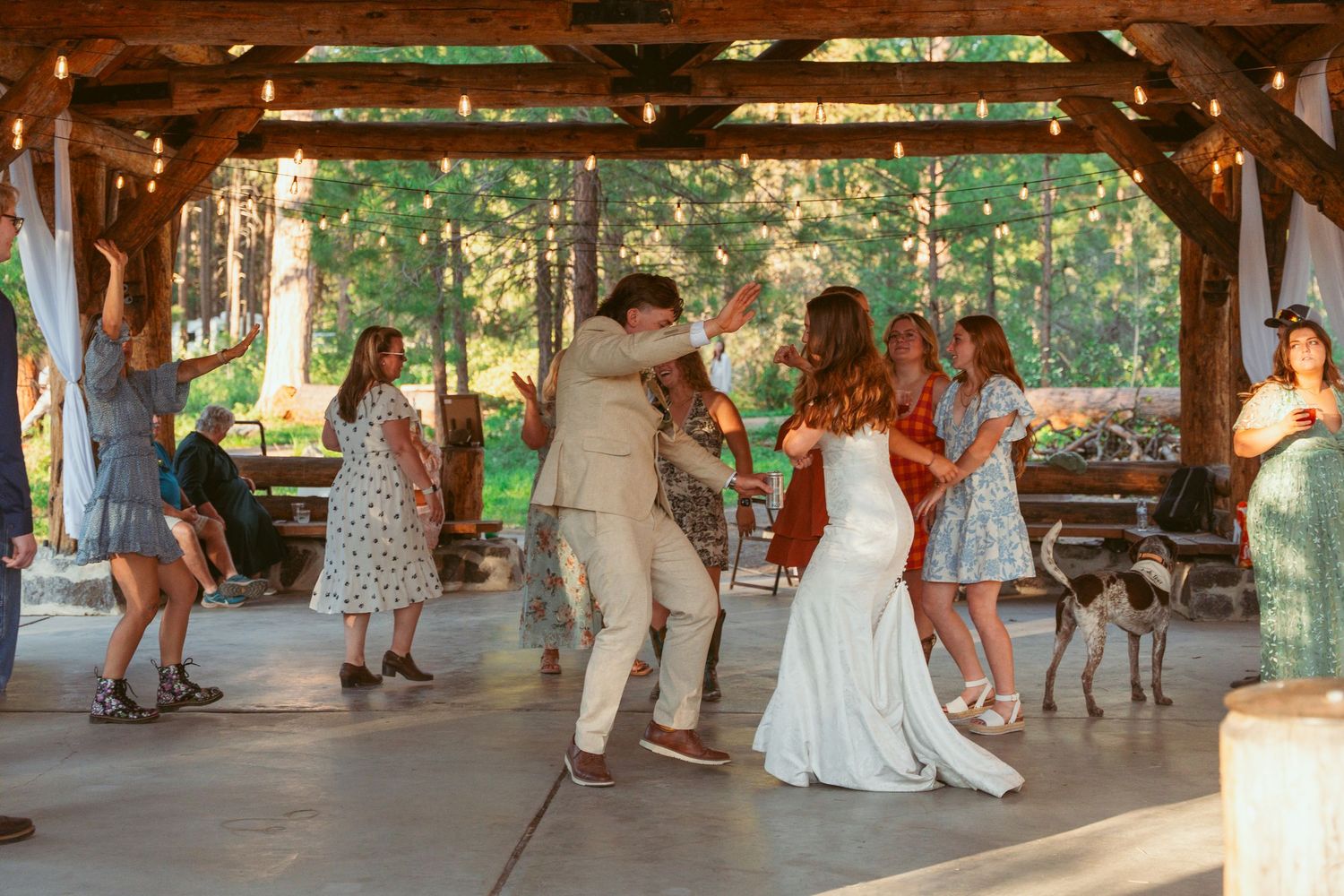Wedding guests dancing under a rustic wooden pavilion decorated with string lights.