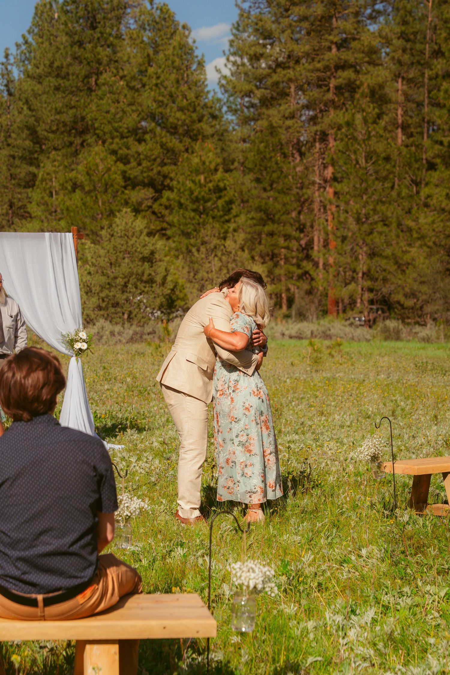 Wedding ceremony setup in a meadow with wooden benches and white fabric decor.