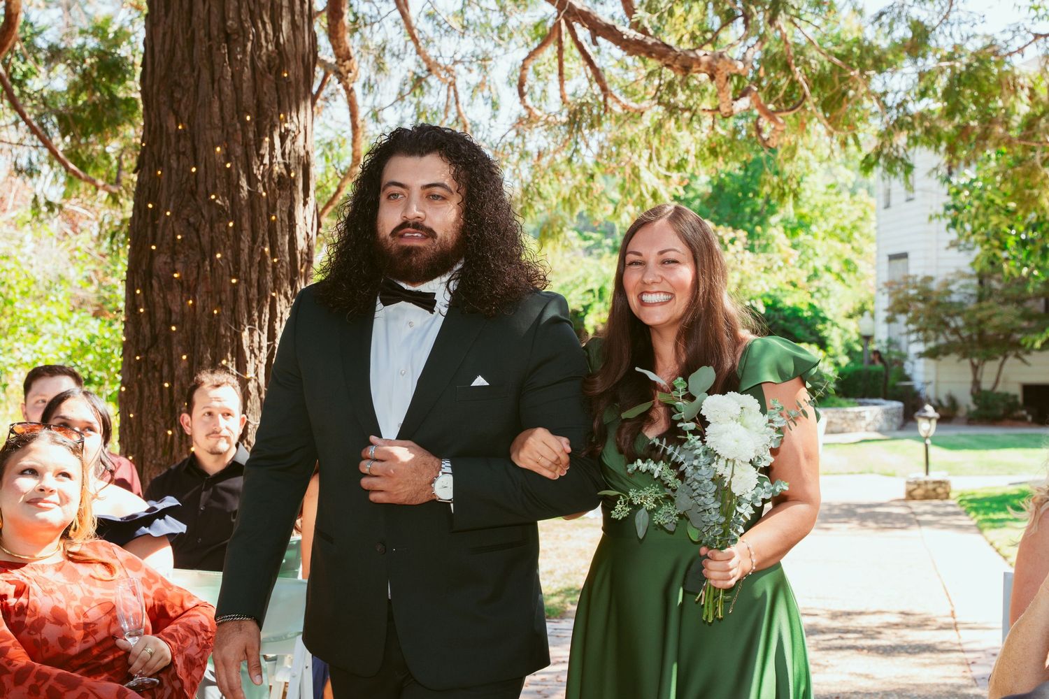 Wedding party members walk down the aisle during an outdoor ceremony beneath towering trees.