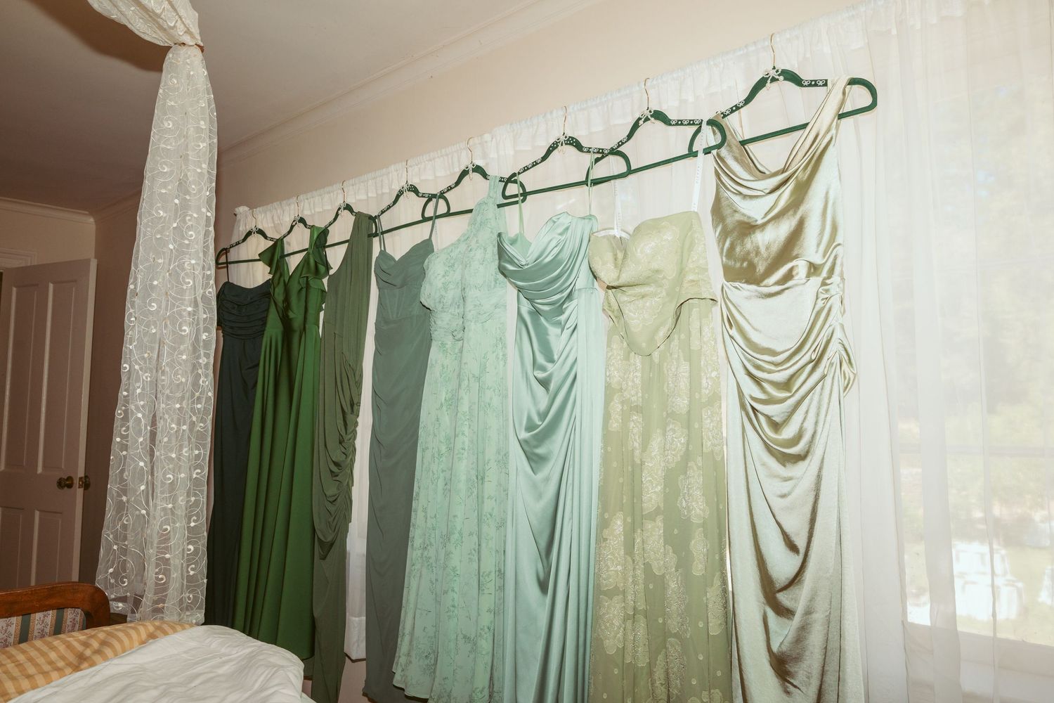 Collection of floor-length dresses in sage, mint and emerald tones displayed on hangers against a wall.