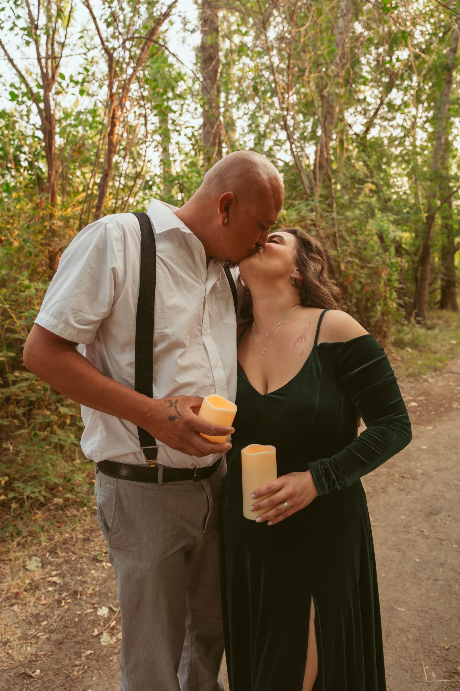 A romantic couple shares an intimate moment on a wooded path with glowing candles at sunset.