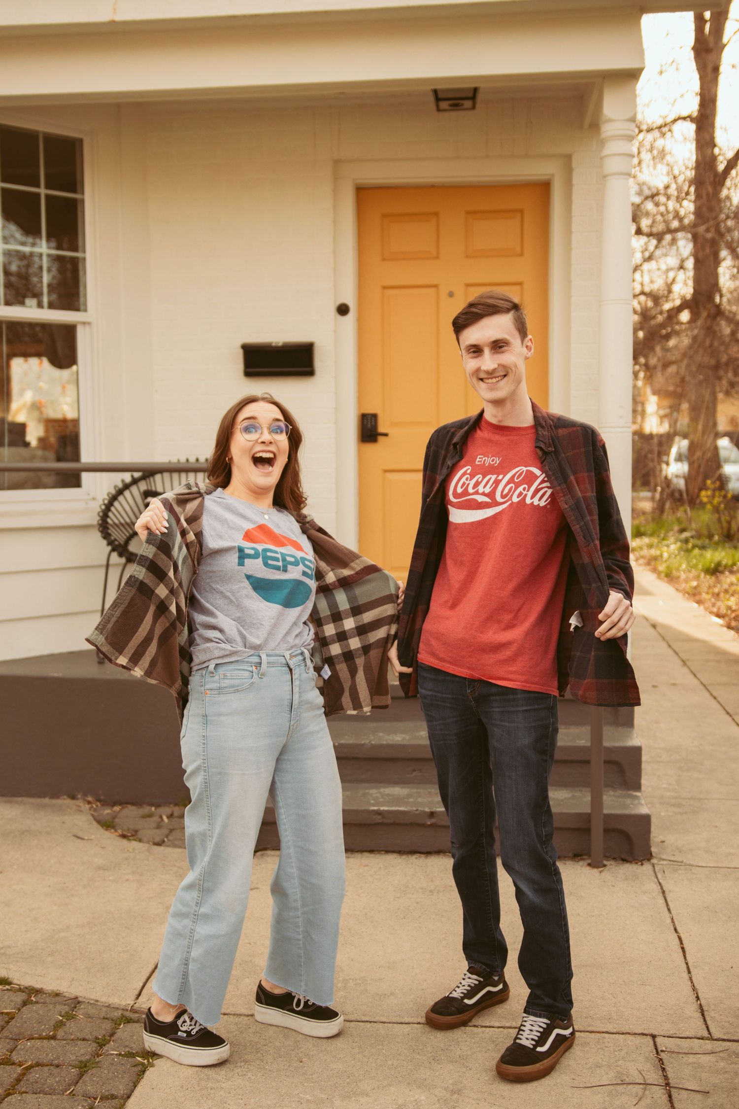 A playful couple wearing Pepsi and Coca-Cola t-shirts pose in front of a house with a yellow door.