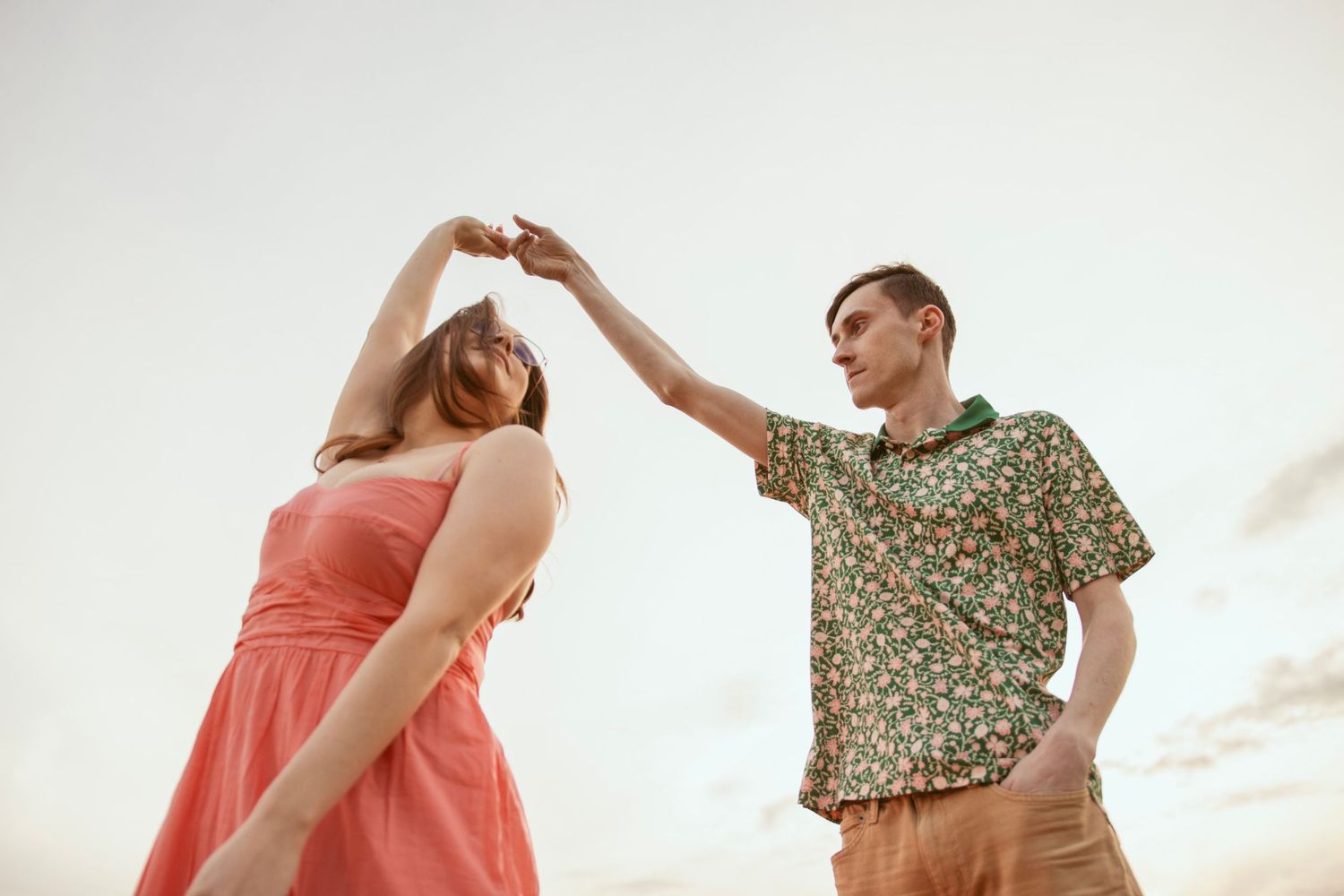 Dancing couple twirls against sunset sky with coral dress and floral shirt in motion.