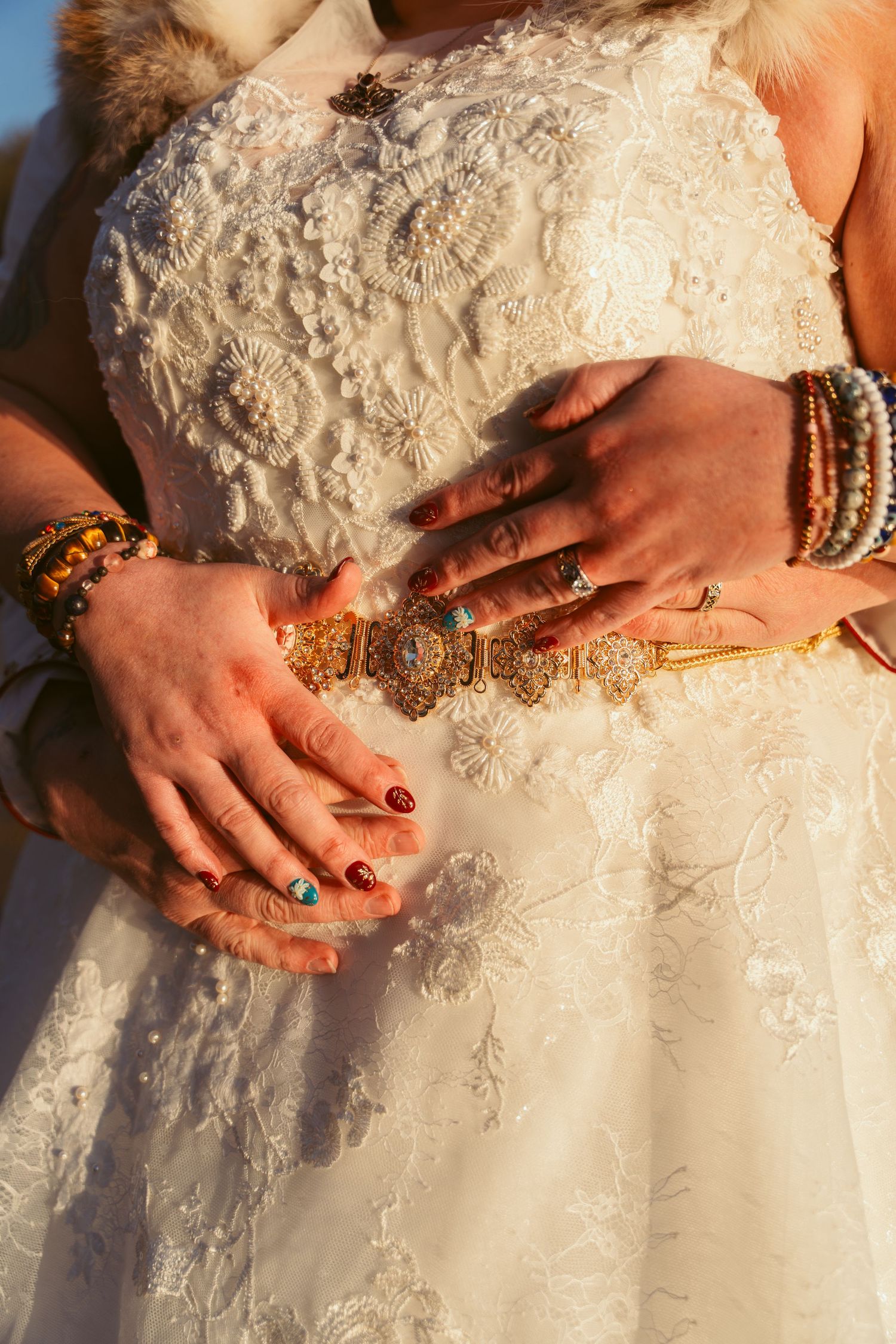 Close-up of hands adorned with bracelets resting on a white lace wedding dress.