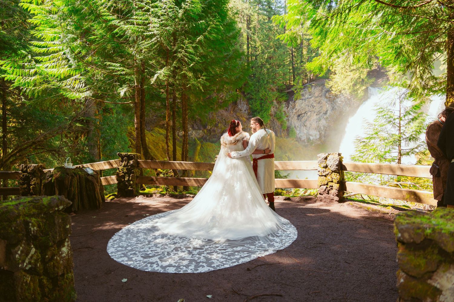 A white wedding dress flows elegantly through a sunlit forest path with tall evergreen trees.