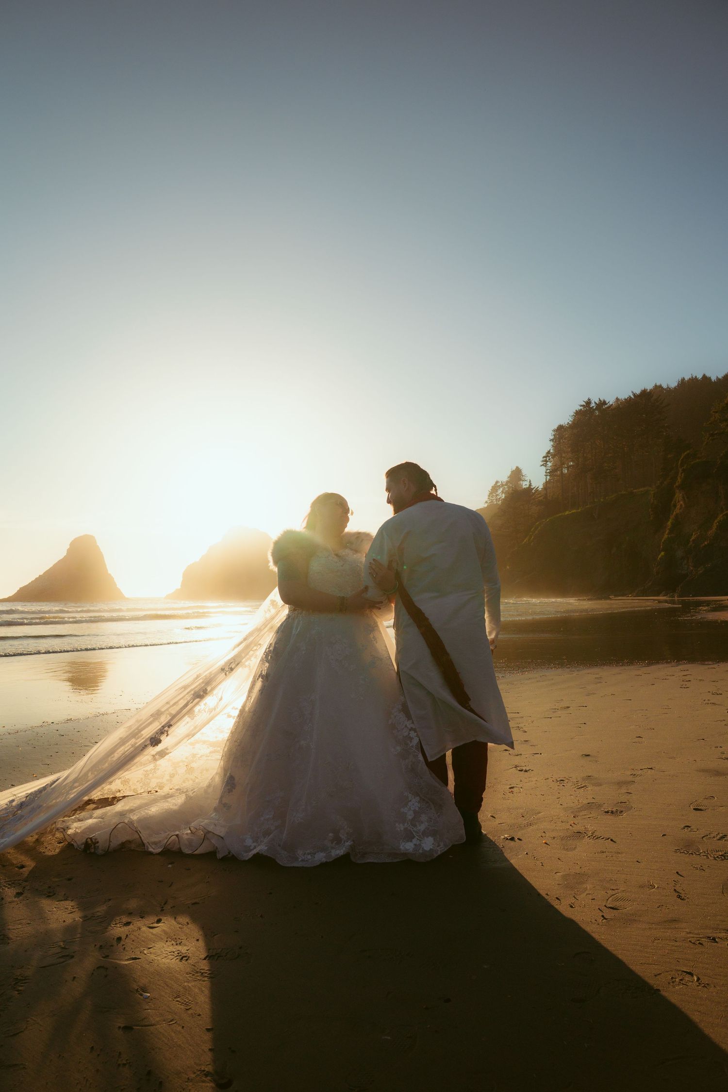 Silhouettes of a couple in wedding attire sit on a beach during golden hour with ocean waves behind them.
