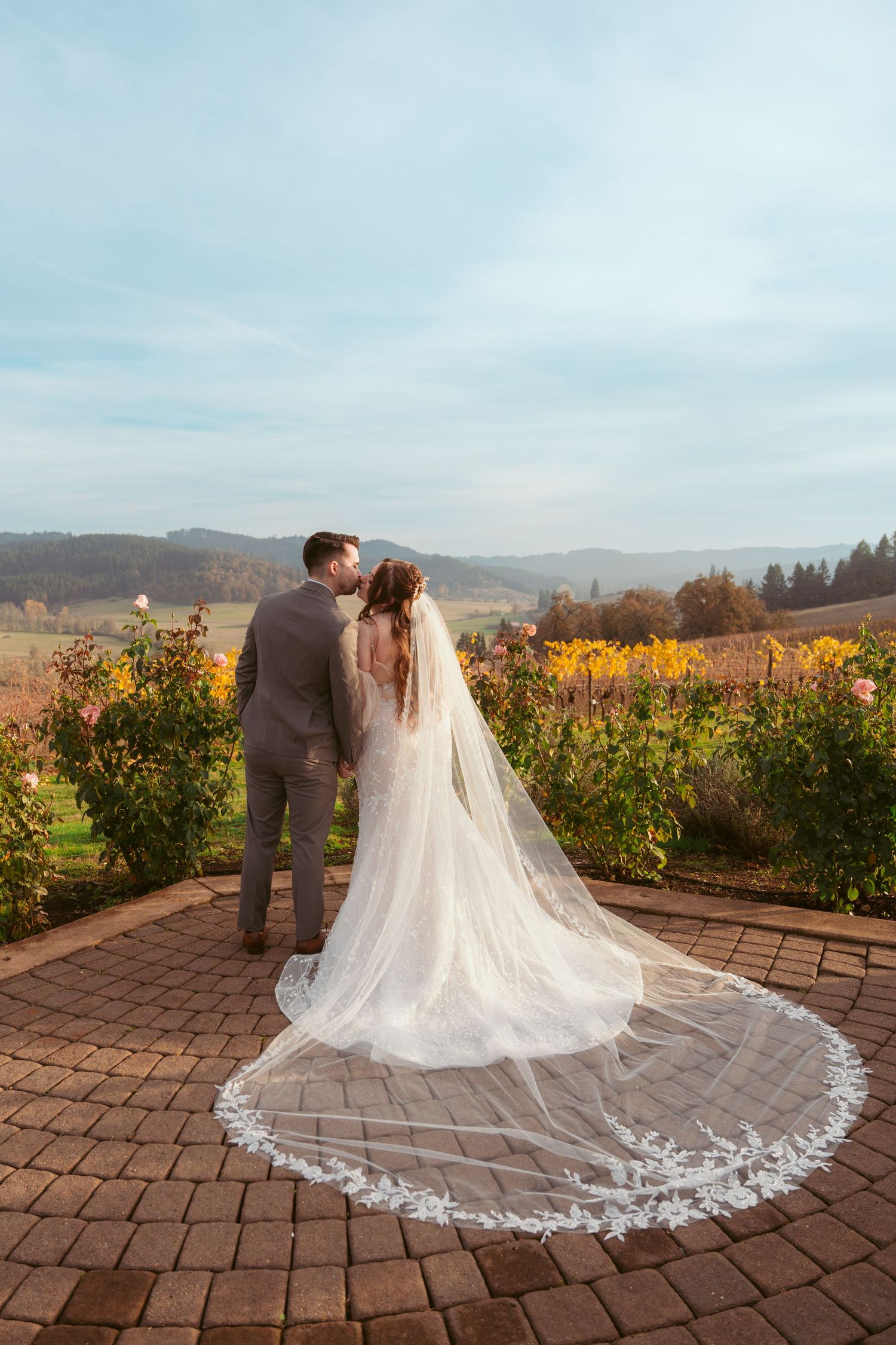 Bride's elegant lace train spreads across brick patio with vineyard views.