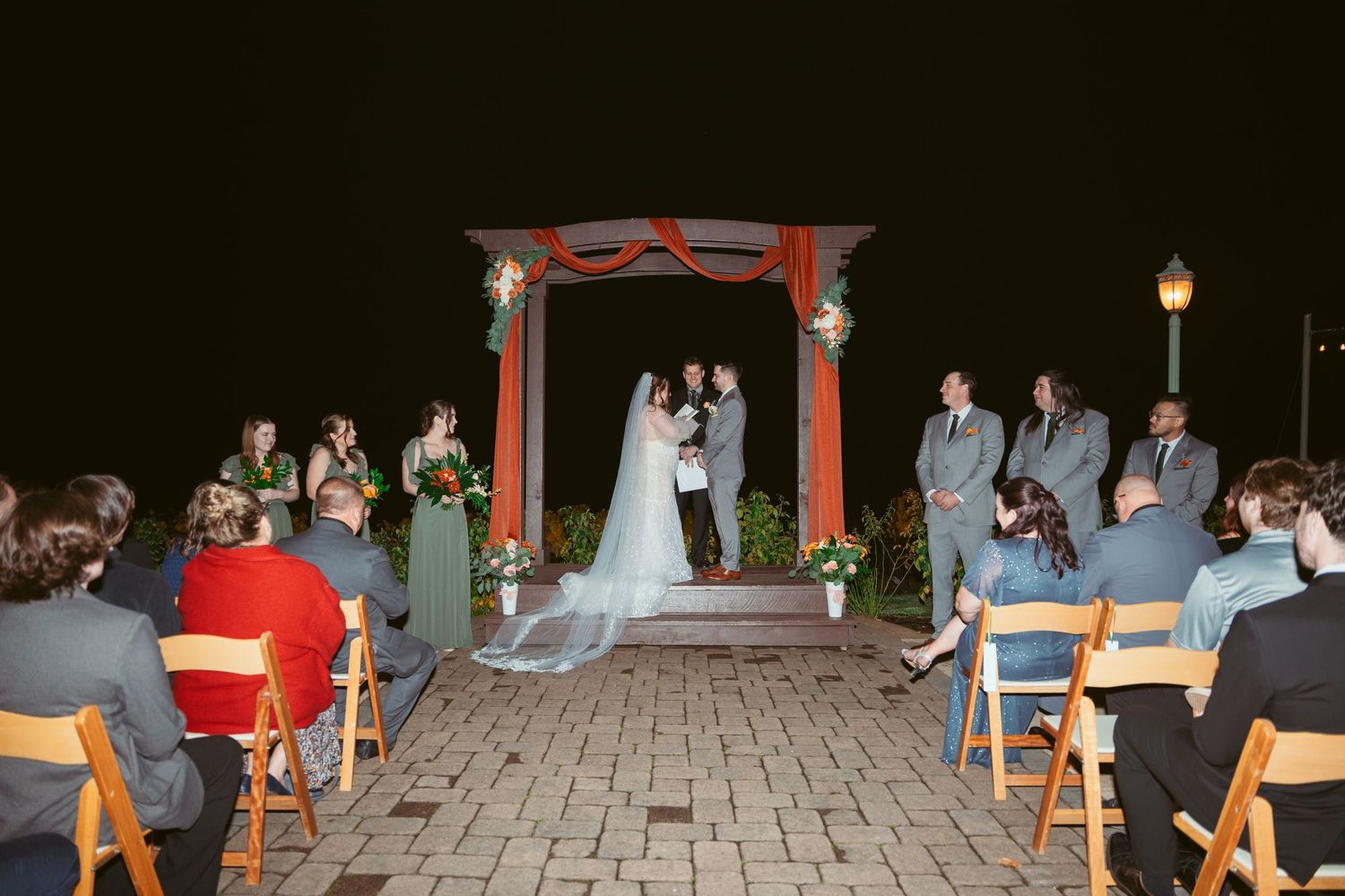 Wide view of an outdoor evening wedding ceremony under a red arch with seated guests.
