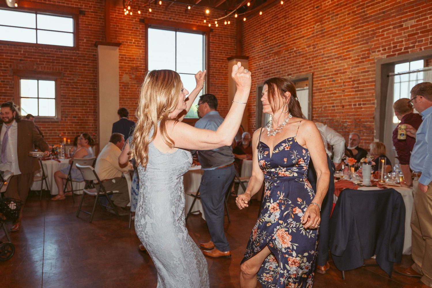Women in formal dresses dance together at a wedding reception in an industrial brick venue.