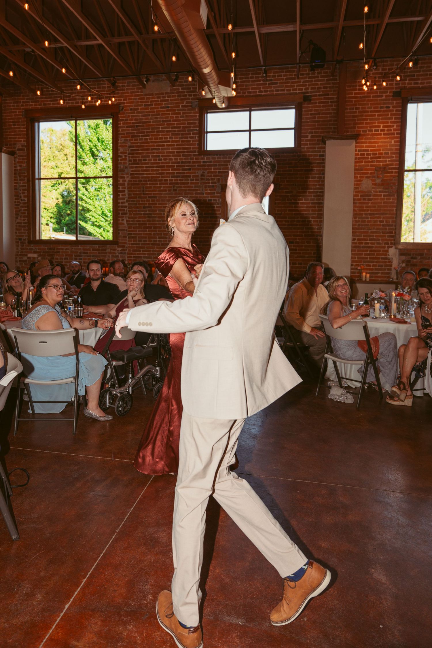 Energetic dance moves on display at a wedding reception in a brick-walled venue with string lights.