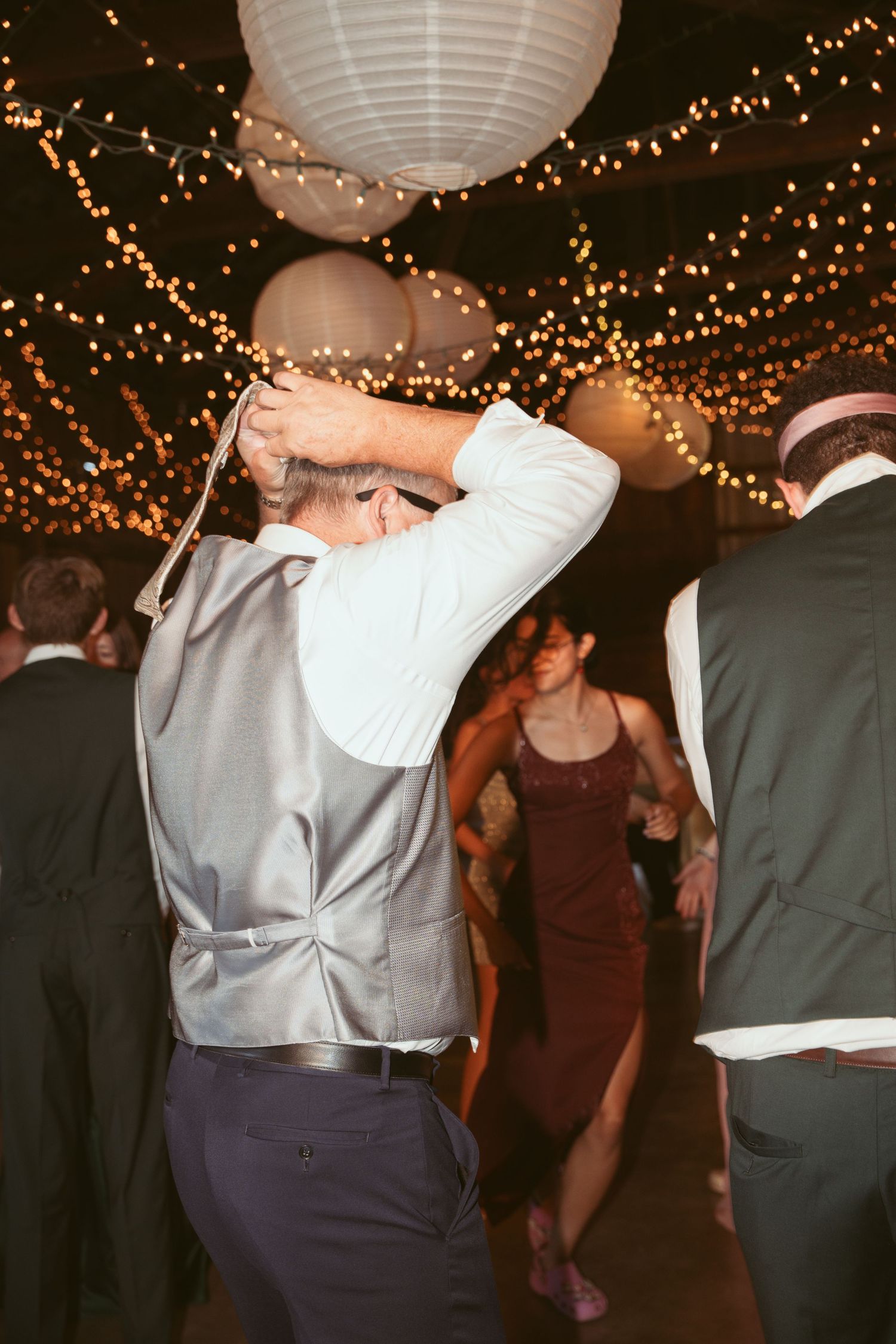 Wedding guests dancing in formal attire under paper lanterns and twinkle lights.