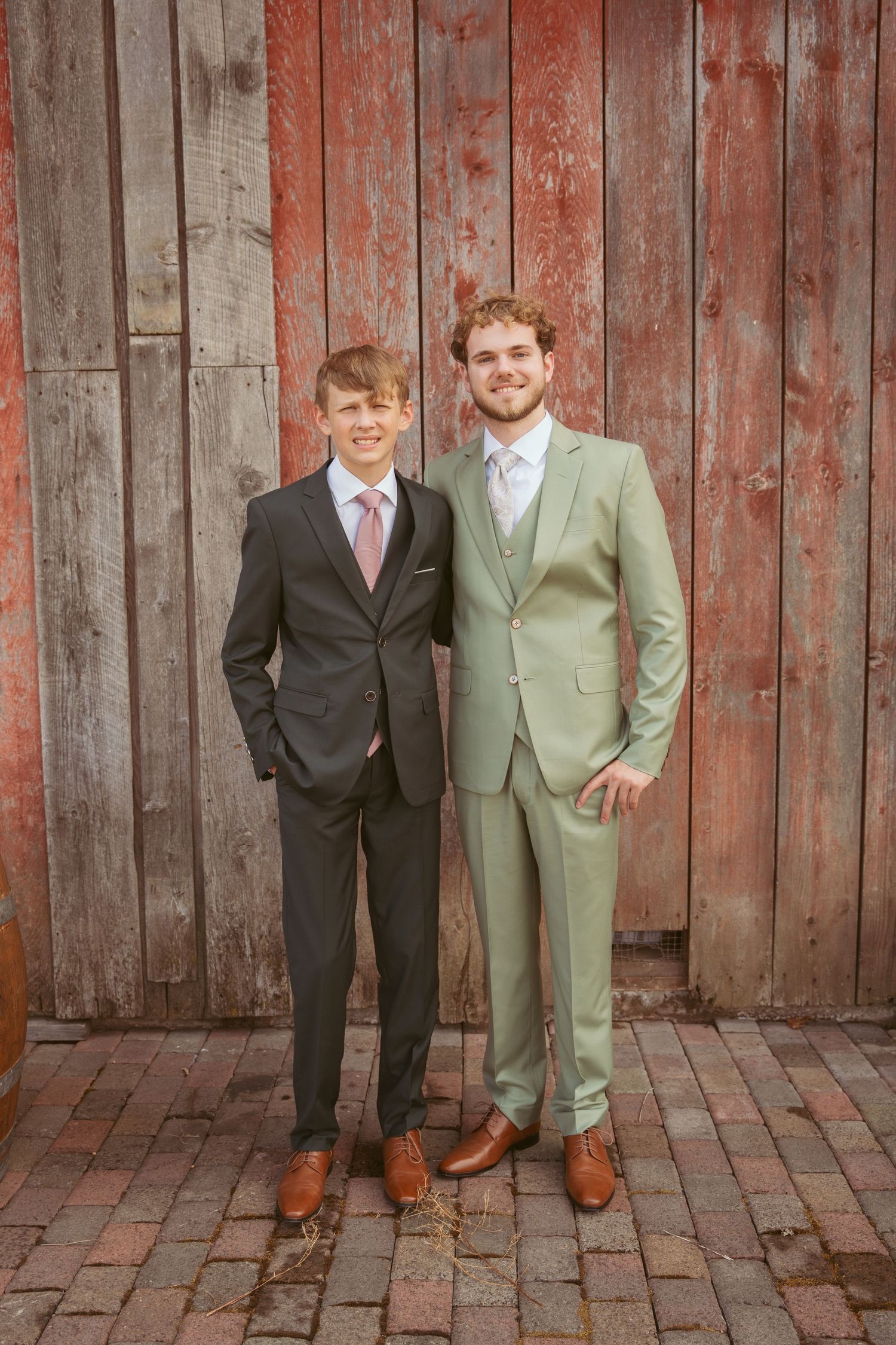 Two people in contrasting suits pose together against weathered red barn wall.