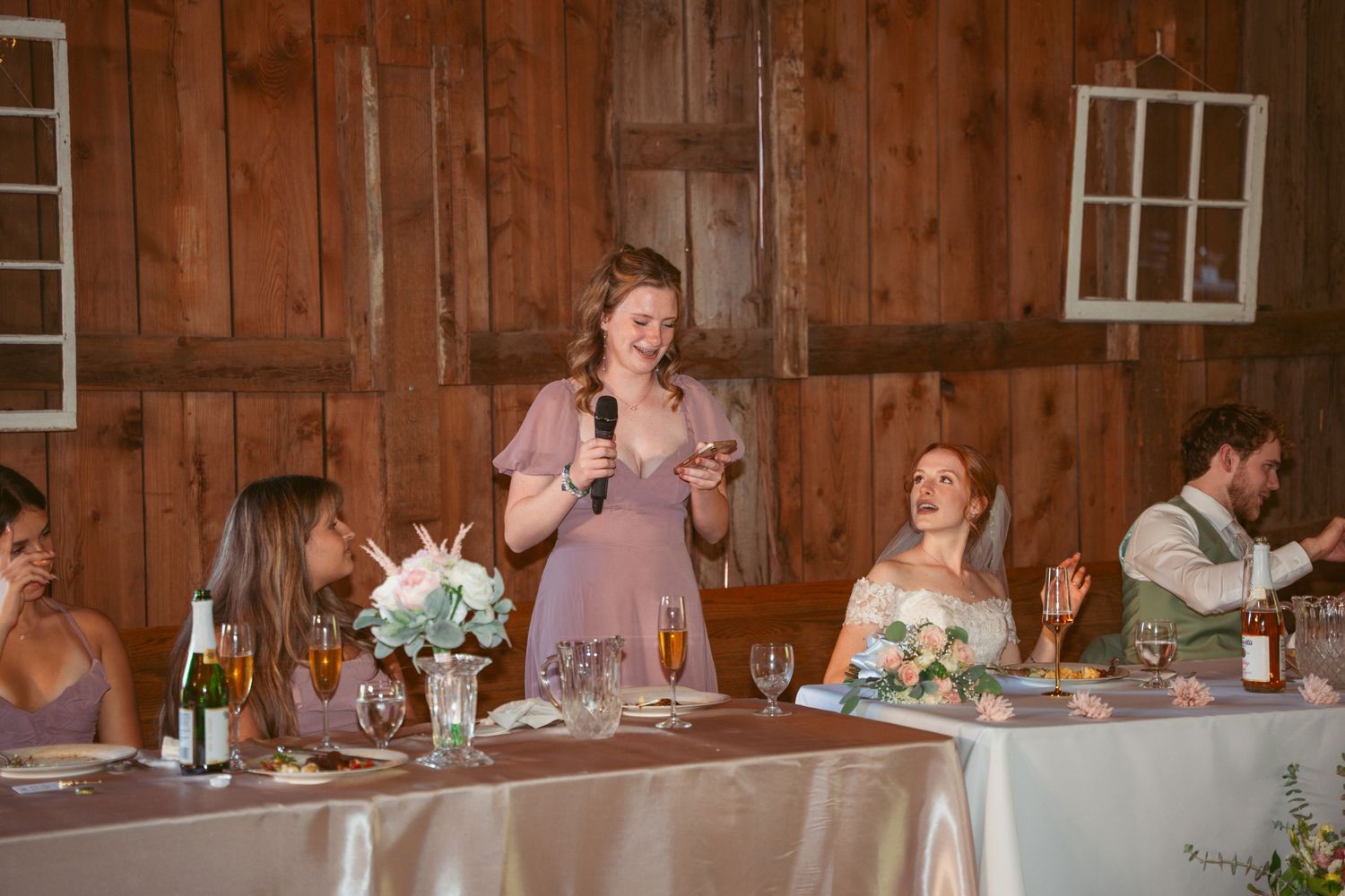 Wedding party dining at decorated head table in rustic barn with wooden paneled walls.