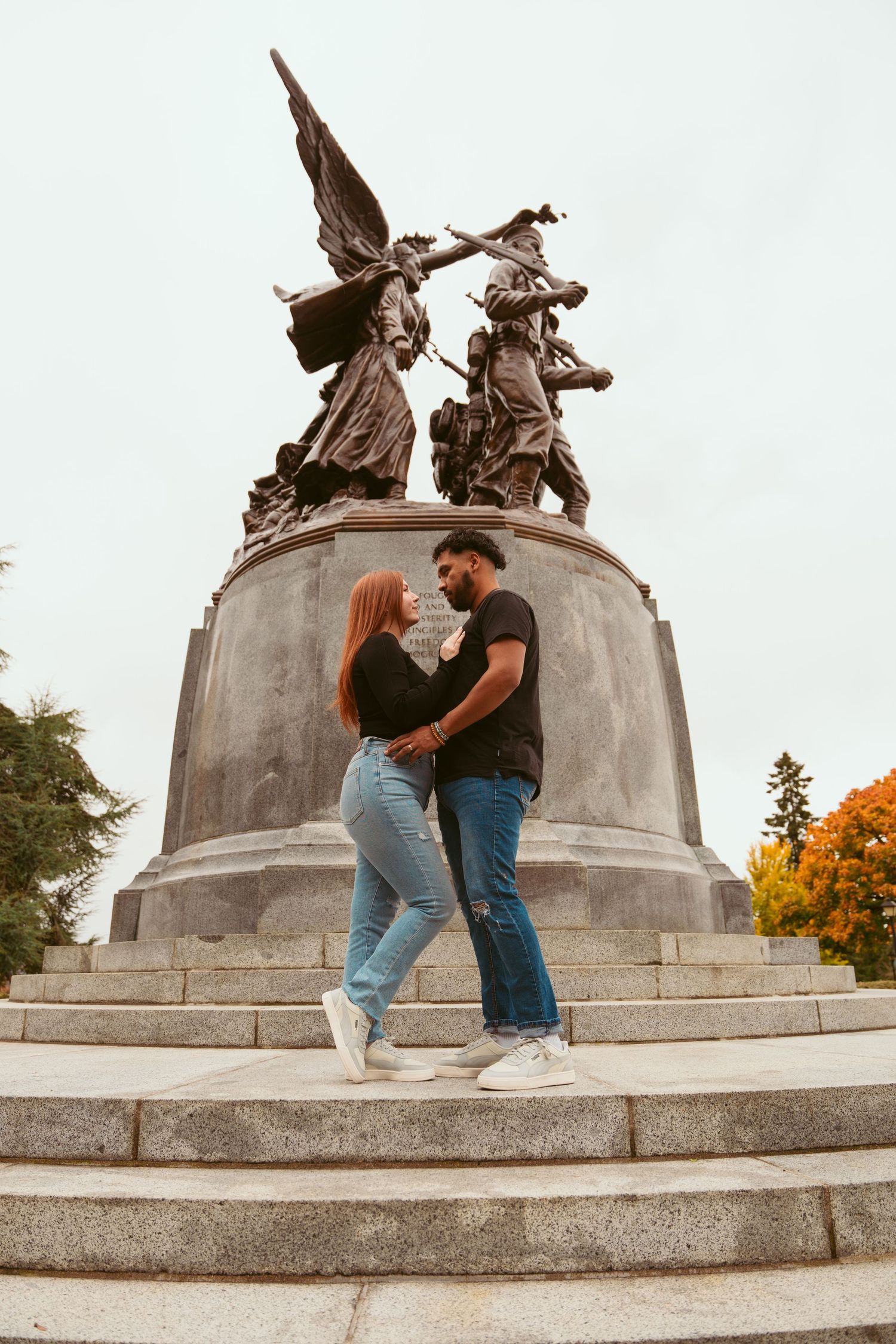 Couple posing playfully near a stone monument with winged statues during autumn.