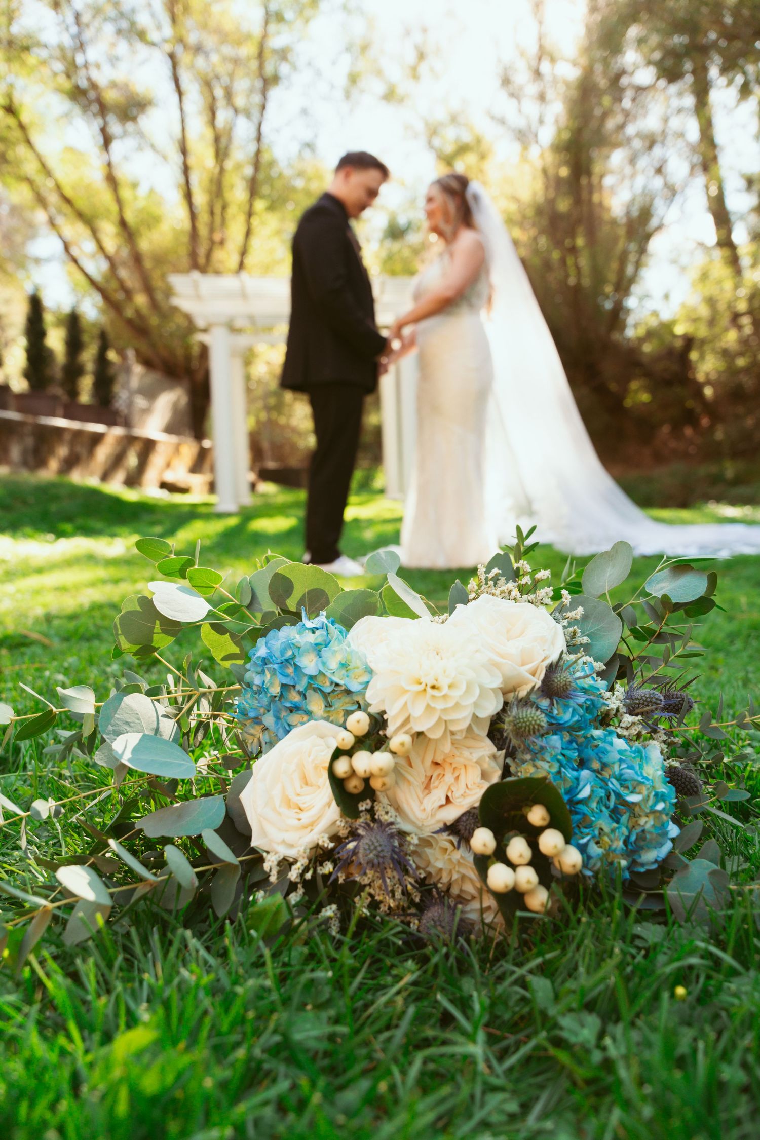An elegant wedding bouquet with white roses and blue hydrangeas rests on green grass.
