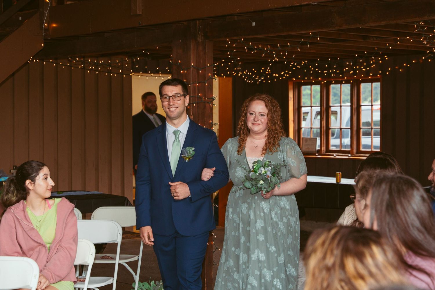 Wedding party members walking down aisle in rustic barn venue with string lights overhead.