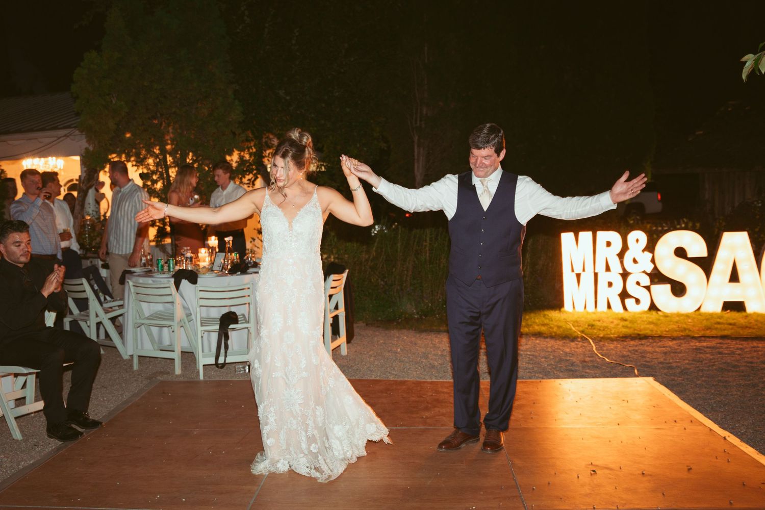 Newlyweds dancing in front of illuminated MR & MRS sign at outdoor reception.