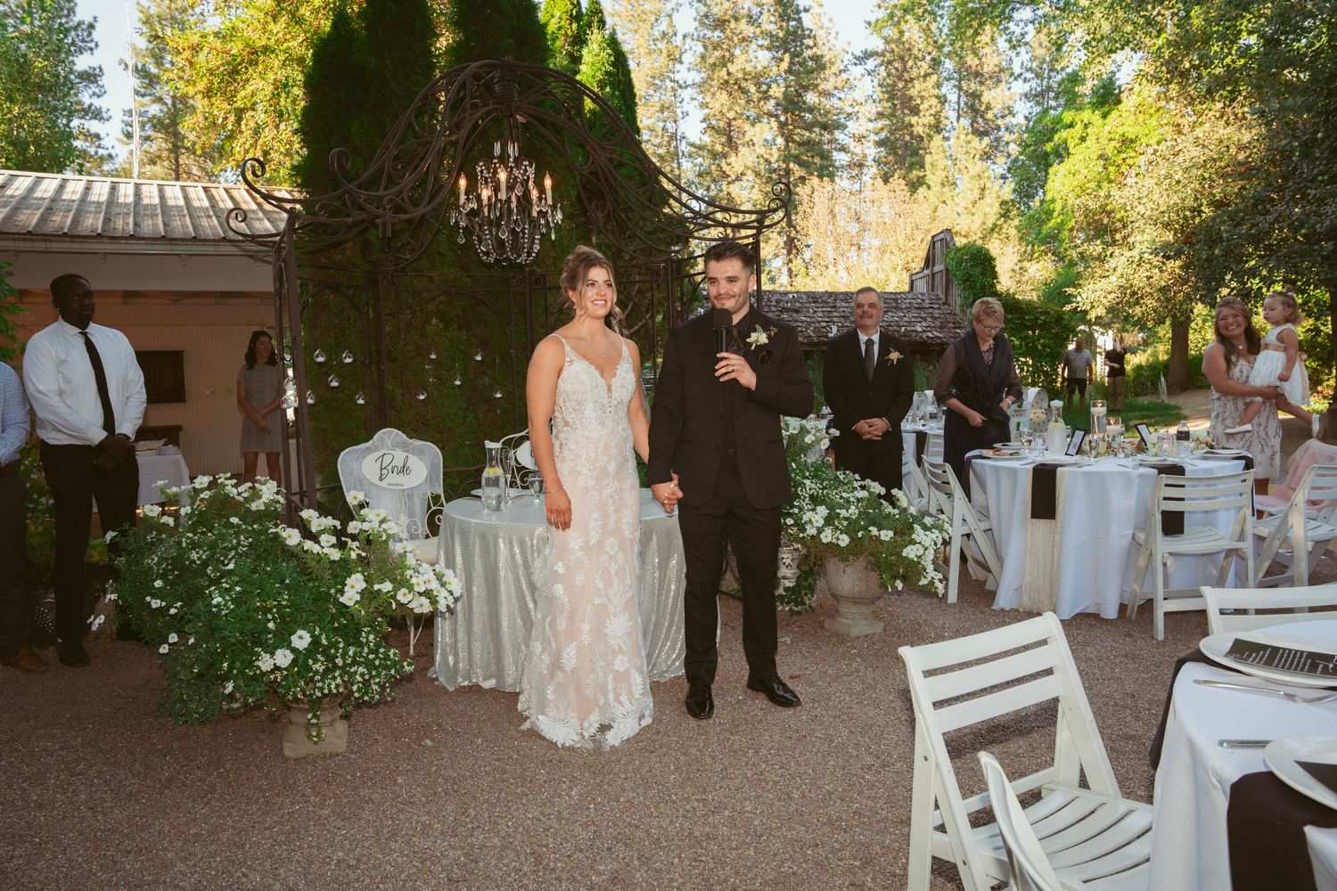 An elegant outdoor wedding reception setup with white chairs, floral arrangements and a rustic wooden structure under tall trees.