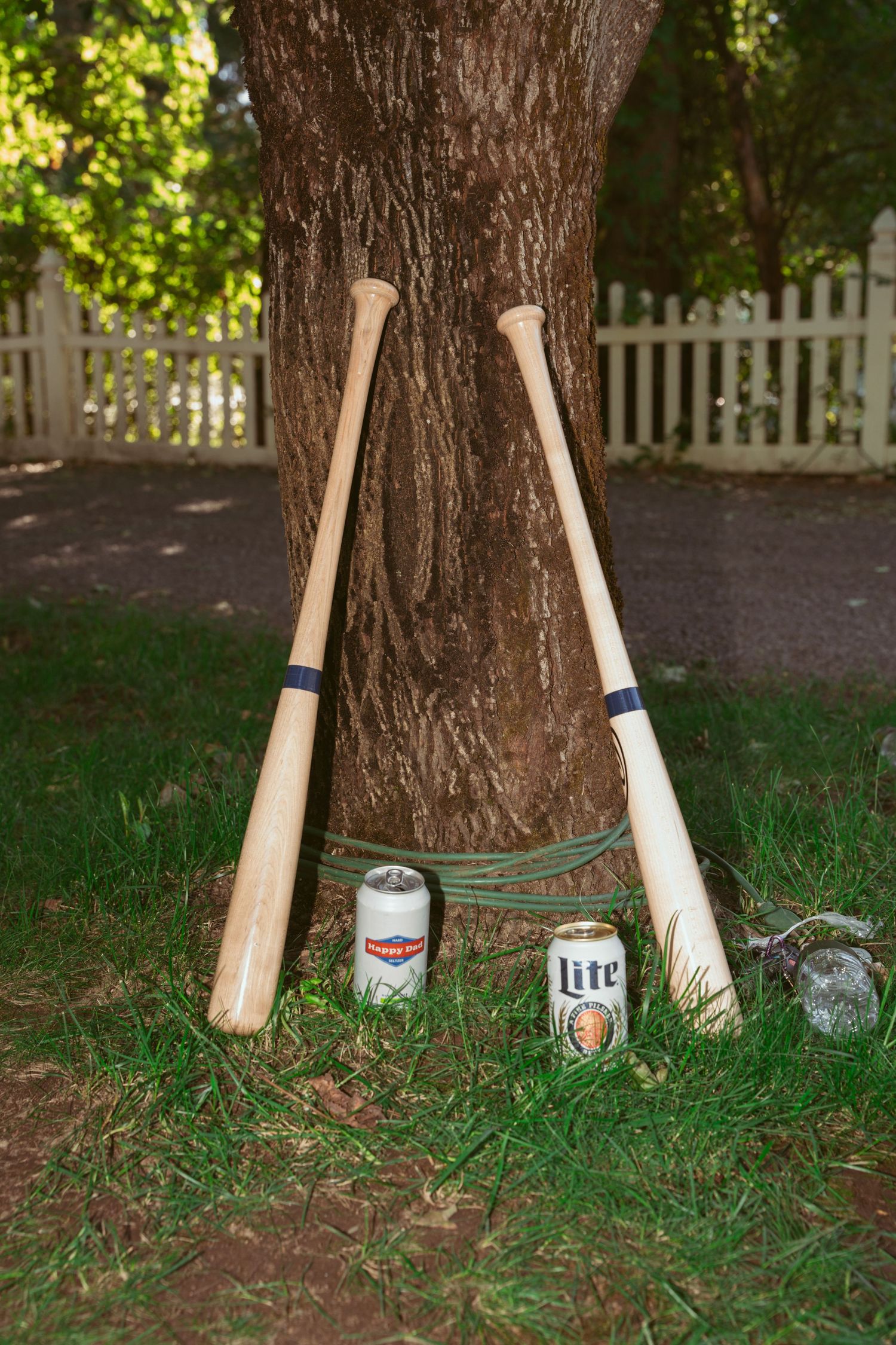 Baseball bats and beer bottles arranged against tree trunks on grass in an artistic display.