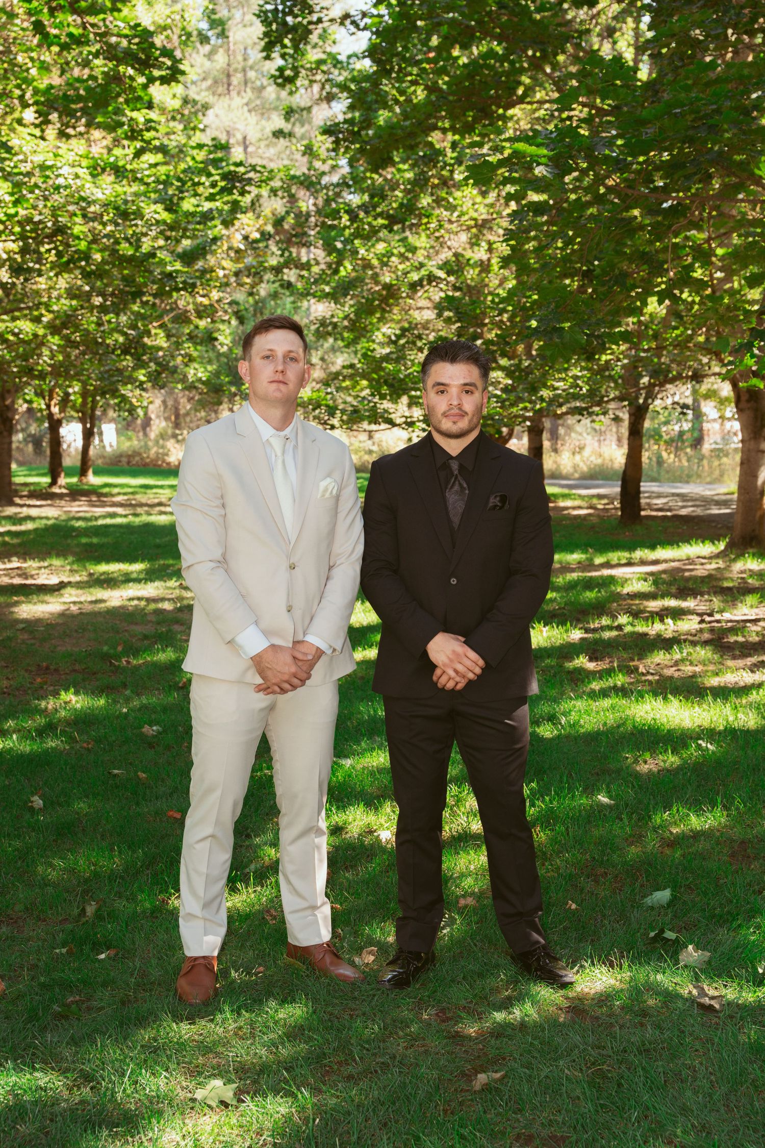 Two people in formal attire stand together on grass under dappled sunlight and trees.
