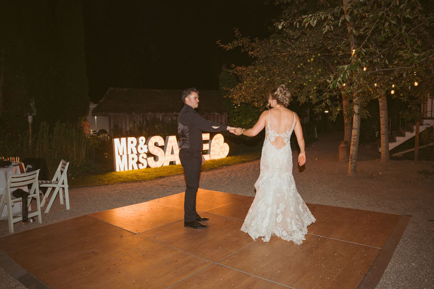 Outdoor wedding reception with illuminated signage and dancing guests at night.