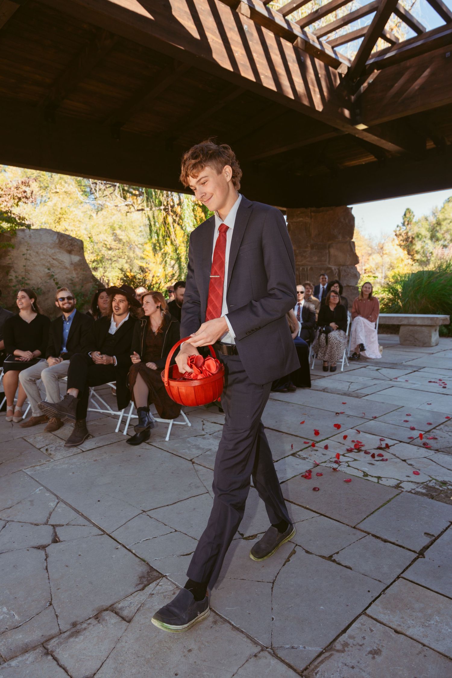 Attendant in dark suit carries red basket while walking down stone pathway during ceremony.
