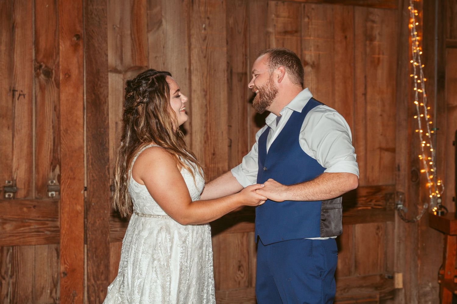 A couple shares a joyful moment while dancing in a barn with string lights.